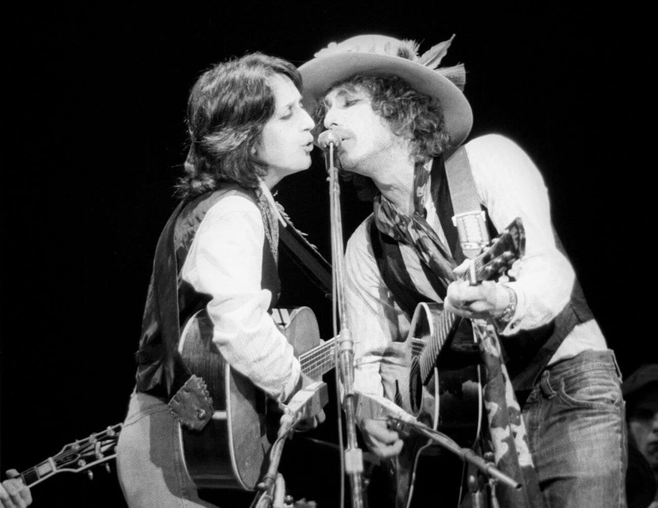 A black and white picture of Bob Dylan and Joan Baez playing guitars and singing into the same microphone.