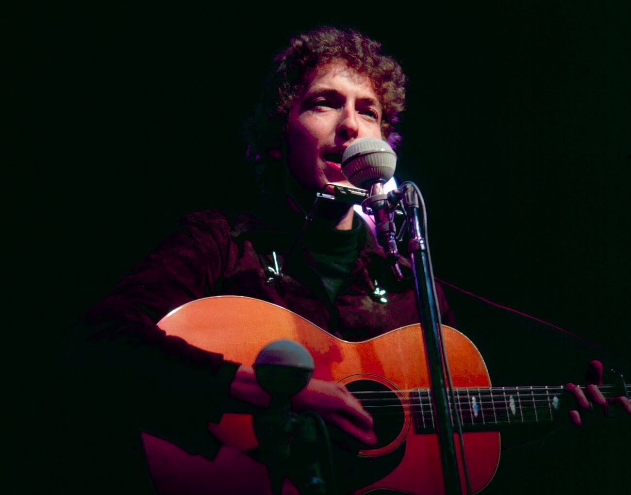 Bob Dylan holds a guitar and sings into a microphone.
