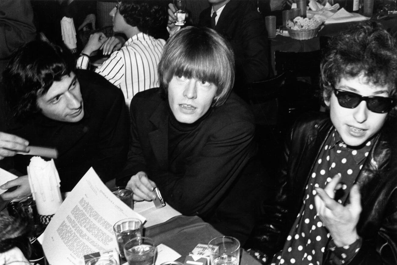 A black and white picture of Brian Jones and Bob Dylan sitting at a table together with a friend.