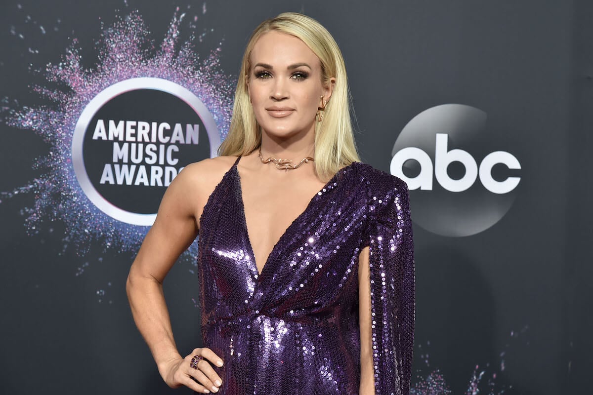 Carrie Underwood poses in a purple gown with one hand on her hip at an event.
