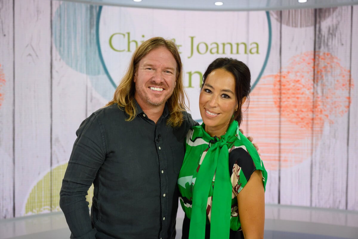 Chip and Joanna Gaines pose in front of a wall with their name on it. Joanna wears a green top and Chip wears a button-down.