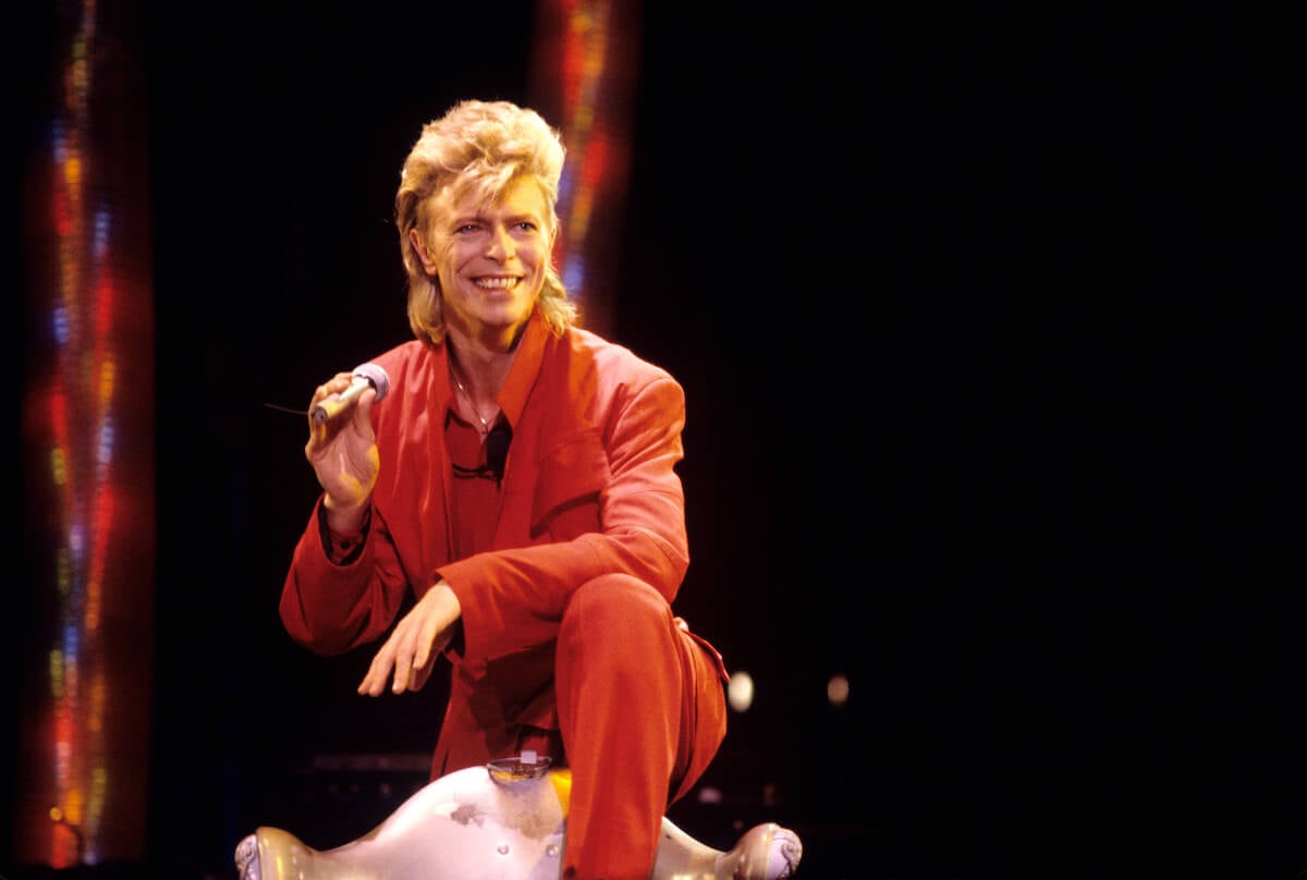 David Bowie crouched down holding a microphone and smiling