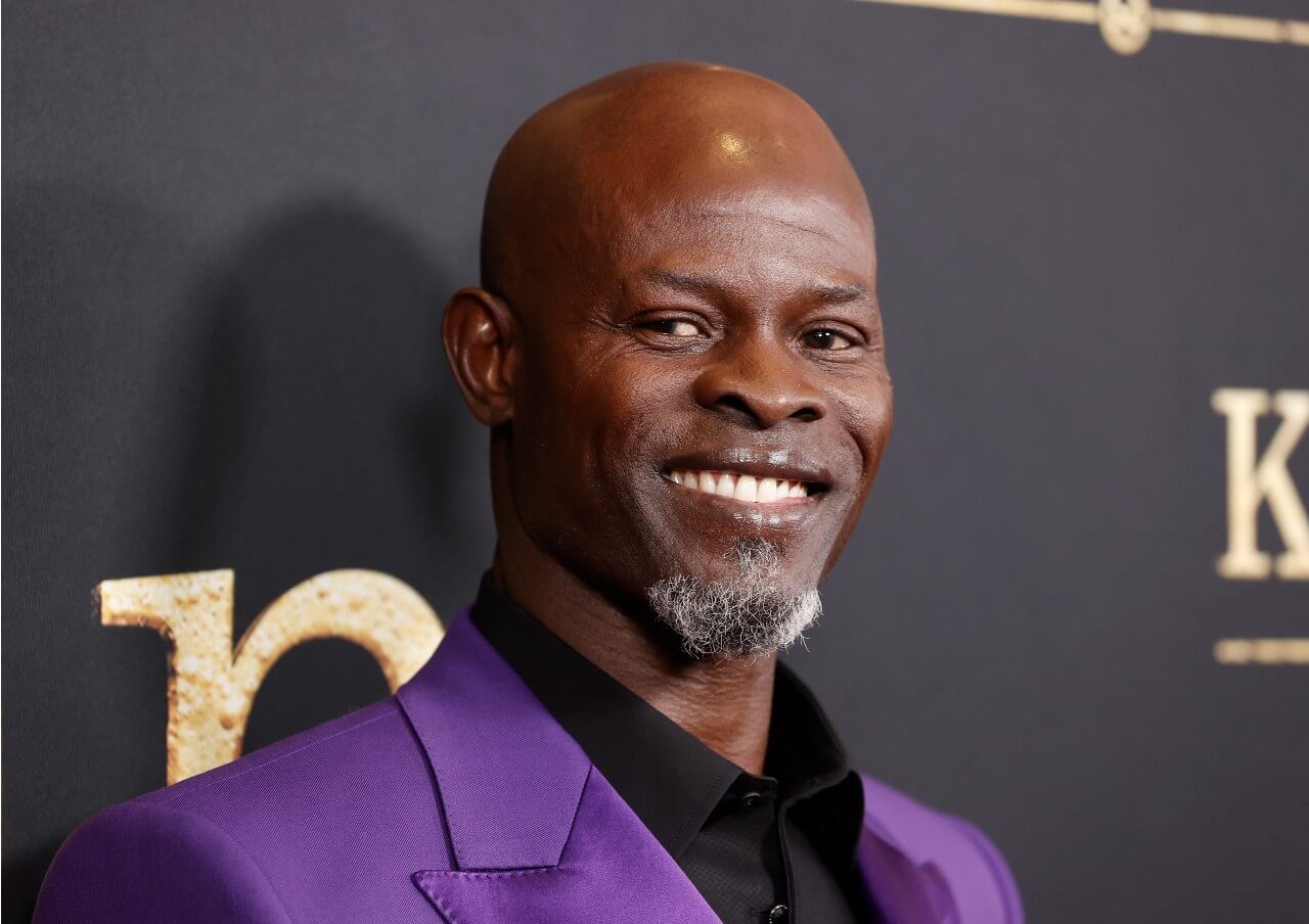 Djimon Hounsou wears a purple jacket and smiles during an event. 