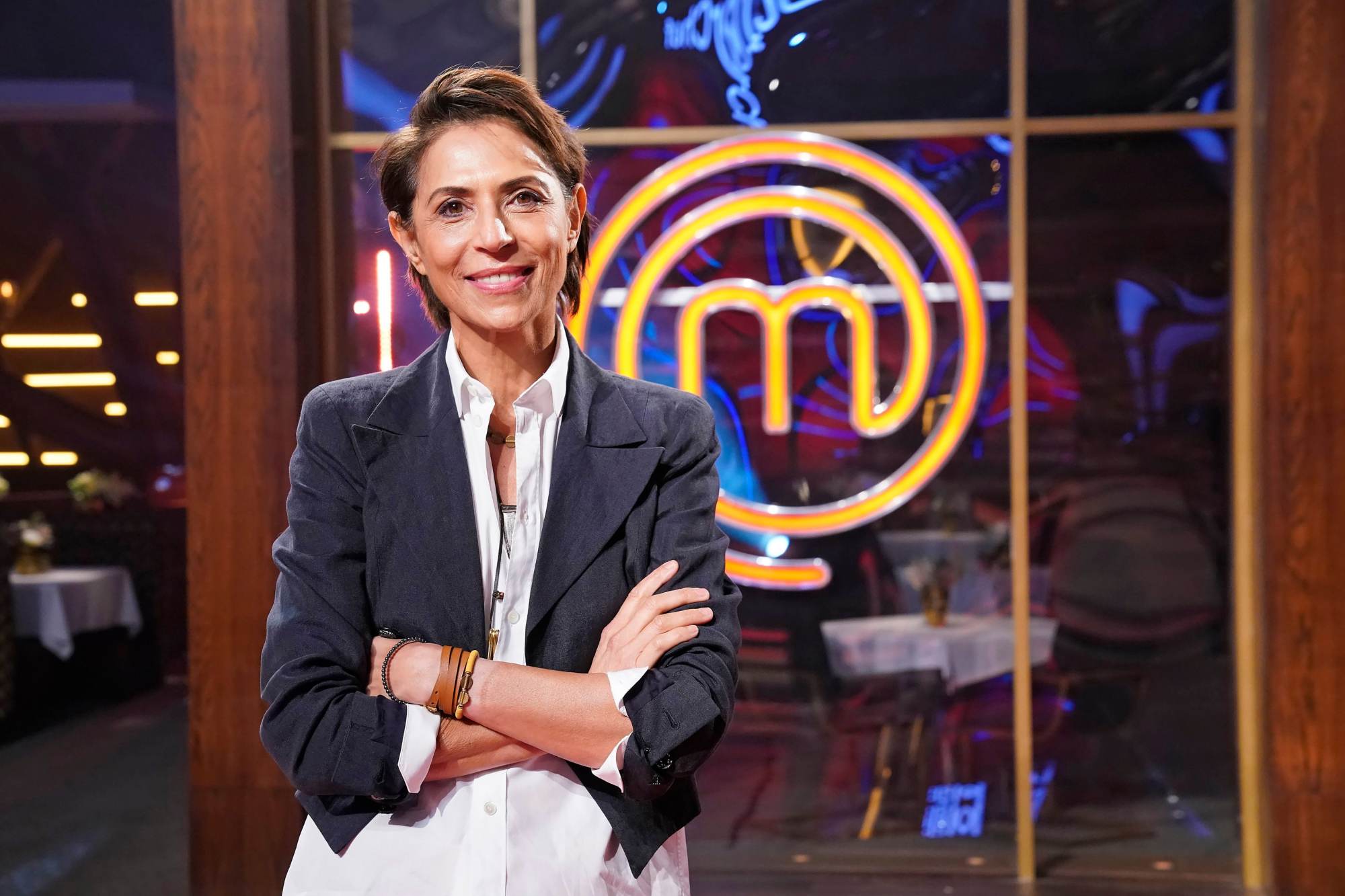 Dominique Crenn standing with her arms crossed in front of the 'MasterChef' logo