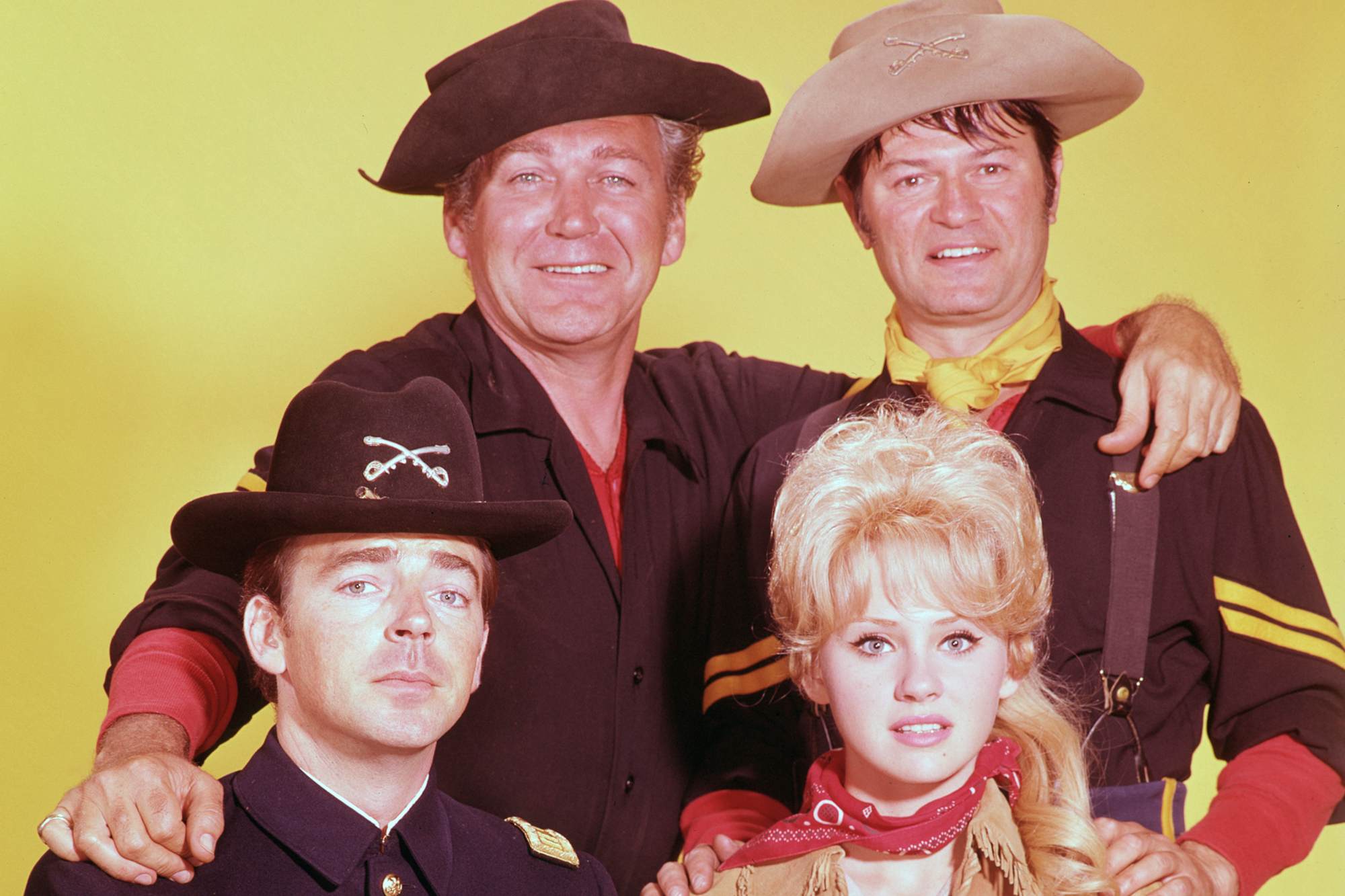 'F Troop' Ken Berry as Captain Wilton Parmenter, Forrest Tucker as Sgt Morgan O'Rourke, Larry Storch as Corporal Randolph Agarn, and Melody Patterson as Wrangler Jane posing in Western costumes in front of a yellow background.