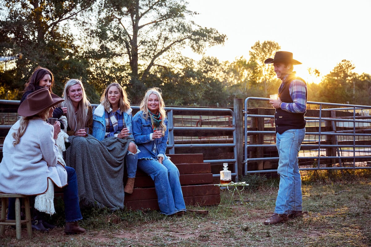 Contestants on 'Farmer Wants a Wife' sitting and smiling while the sun sets and a farmer stands looking at them and smiling