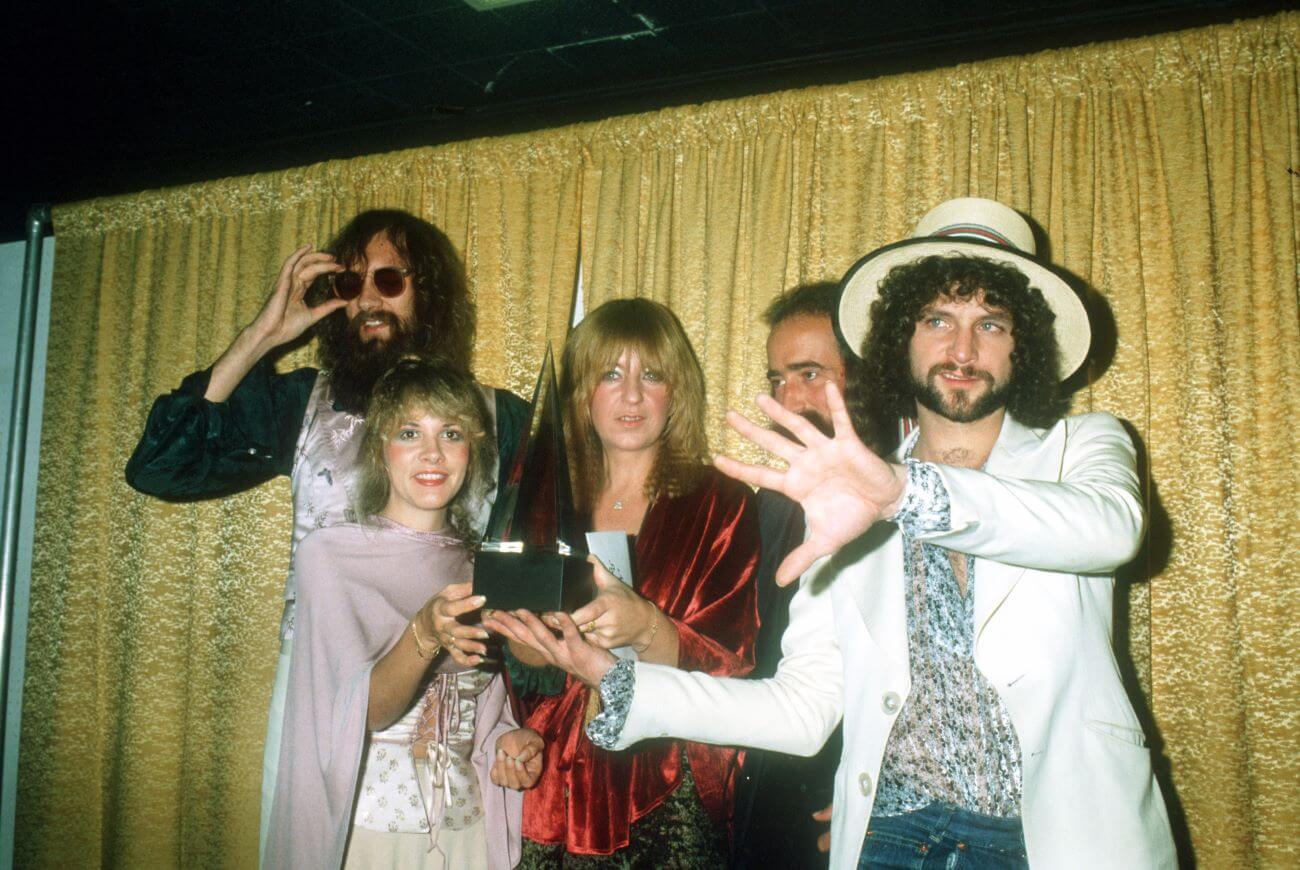 Mick Fleetwood, Stevie Nicks, Christine McVie, John McVie, and Lindsey Buckingham of Fleetwood Mac hold a trophy and stand in front of a gold curtain.
