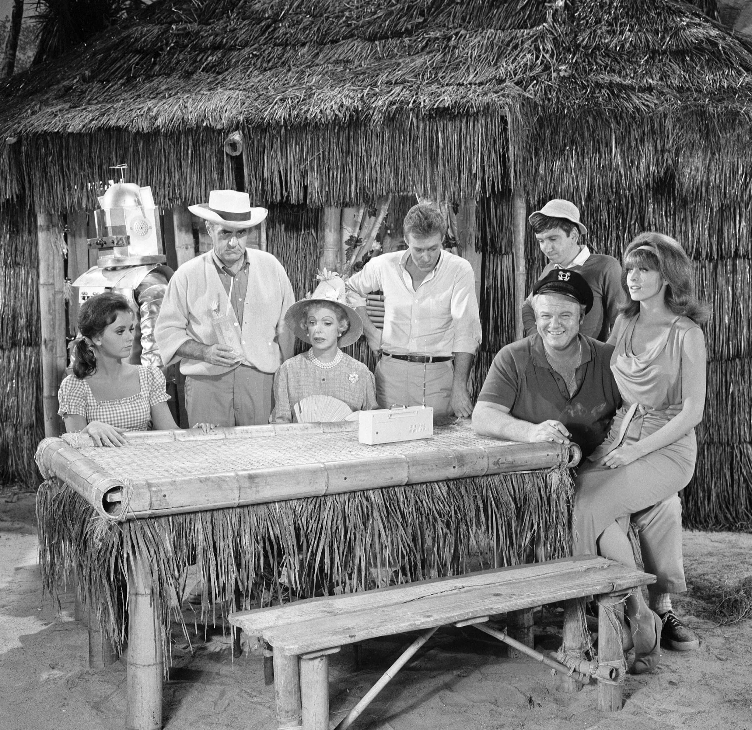 A black and white photo of the 'Gilligan's Island' cast members gathered around a bamboo picnic table on set of the TV comedy.