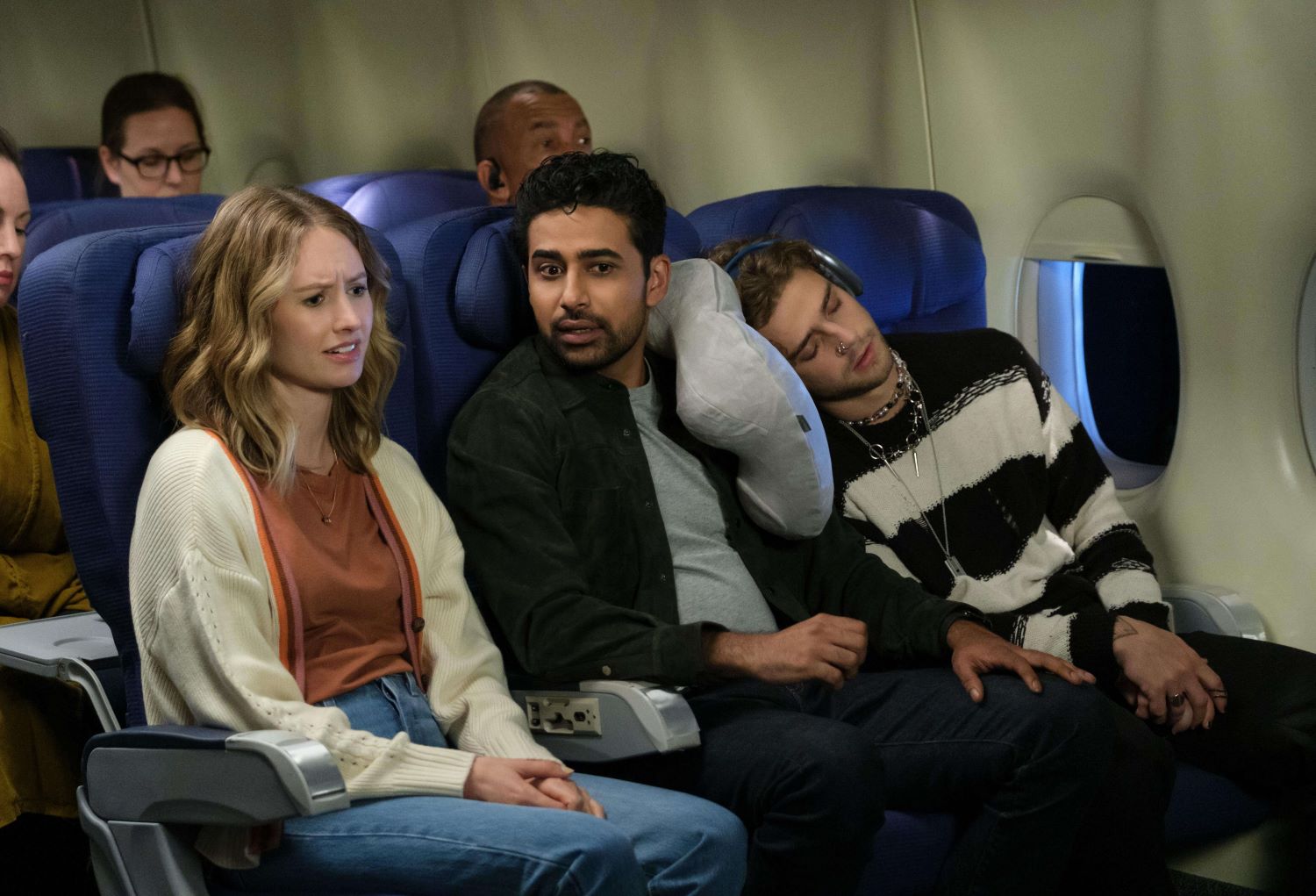 Caitlin Thompson as Taylor and Suraj Sharma as Sid sit next to one another on a plane in 'How I Met Your Father' Season 2 Episode 11, 'Daddy.' Taylor wears a white cardigan over a dark orange shirt and jeans. Sid wear a black button-up shirt over a light gray shirt and black pants.