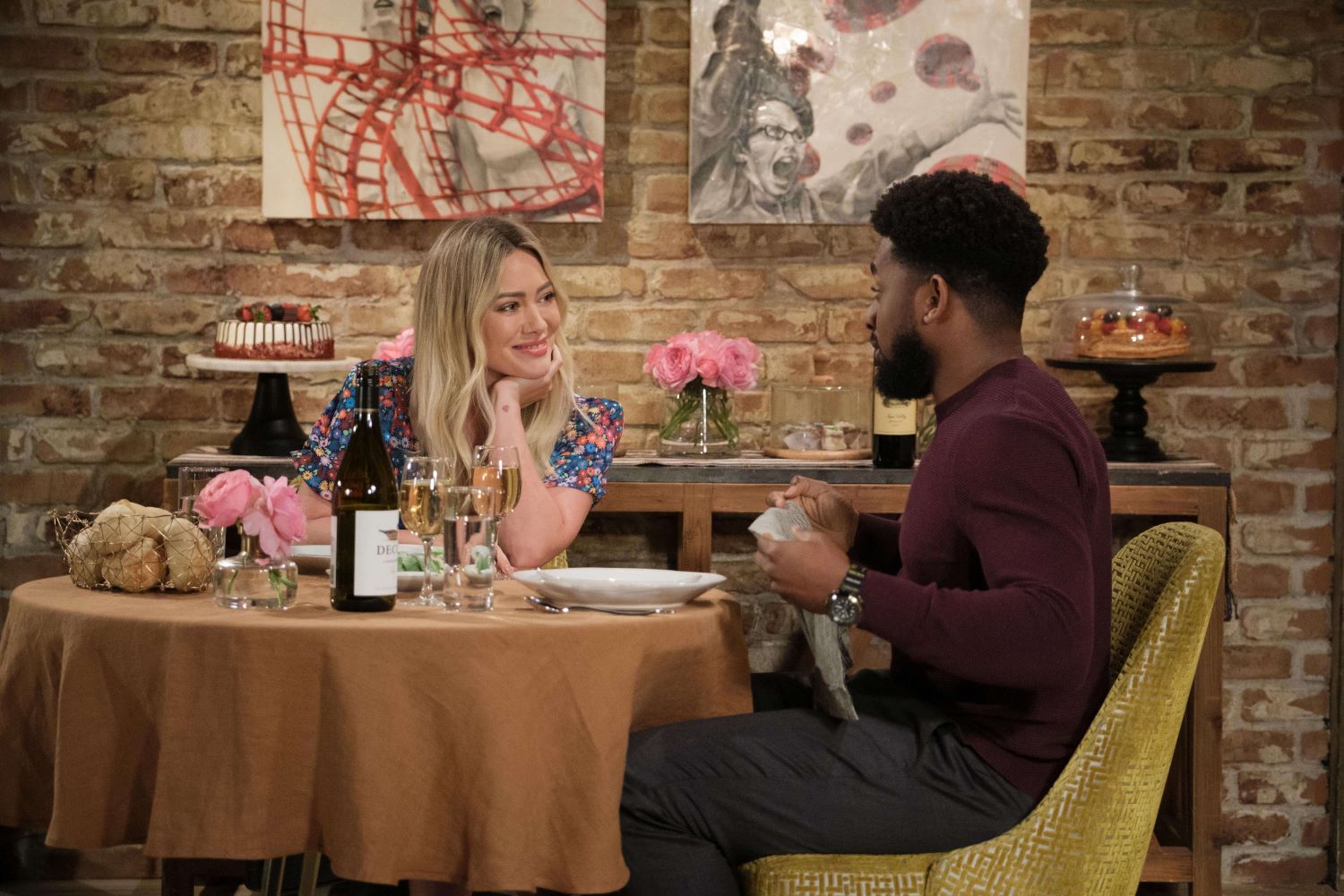 Hilary Duff as Sophie and Daniel Augustin as Ian eat dinner at a restaurant in 'How I Met Your Father' Season 2 Episode 7 on Hulu. Sophie wears a multi-colored floral dress. Ian wears a maroon sweater and dark gray pants.
