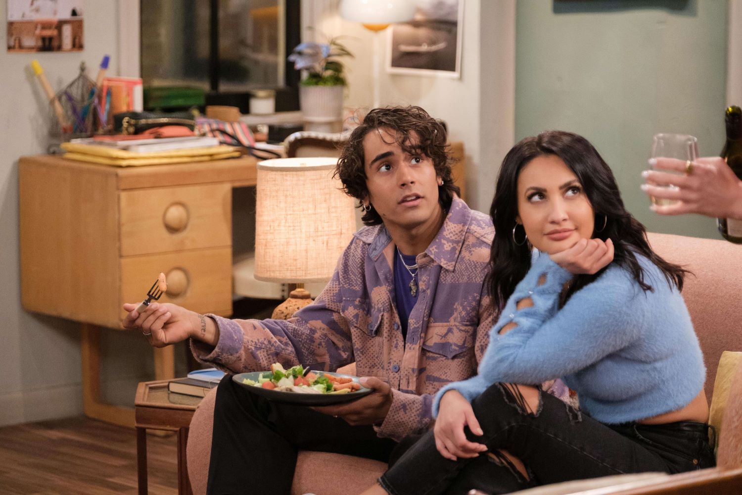 Michael Cimino as Swish and Francia Raisa as Valentina sit on a couch in 'How I Met Your Father' Season 2 Episode 10, 'I'm His Swish.' Swish wears a light purple and orange button-up shirt over a dark purple shirt and black pants. Valentine wears a light blue fuzzy sweater and black ripped jeans.
