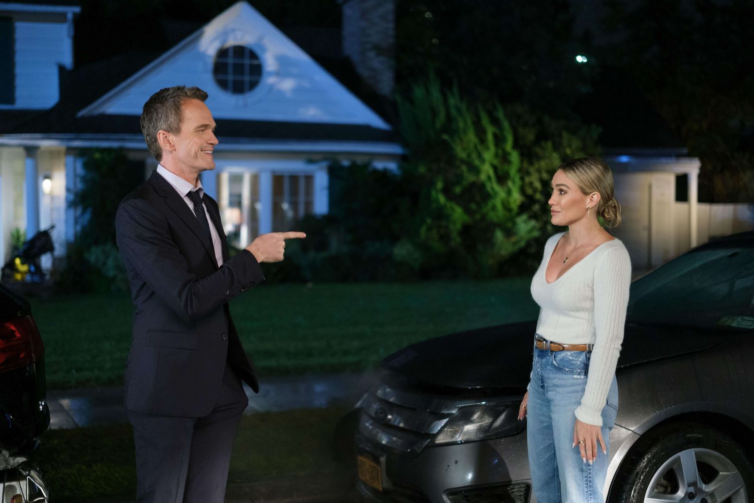 Neil Patrick Harris as Barney Stinson and Hilary Duff as Sophie Tompkins in 'How I Met Your Father' Season 2 on Hulu. Barney wears a black suit over a white button-up shirt and black tie. Sophie wears a white long-sleeved shirt and light blue jeans.