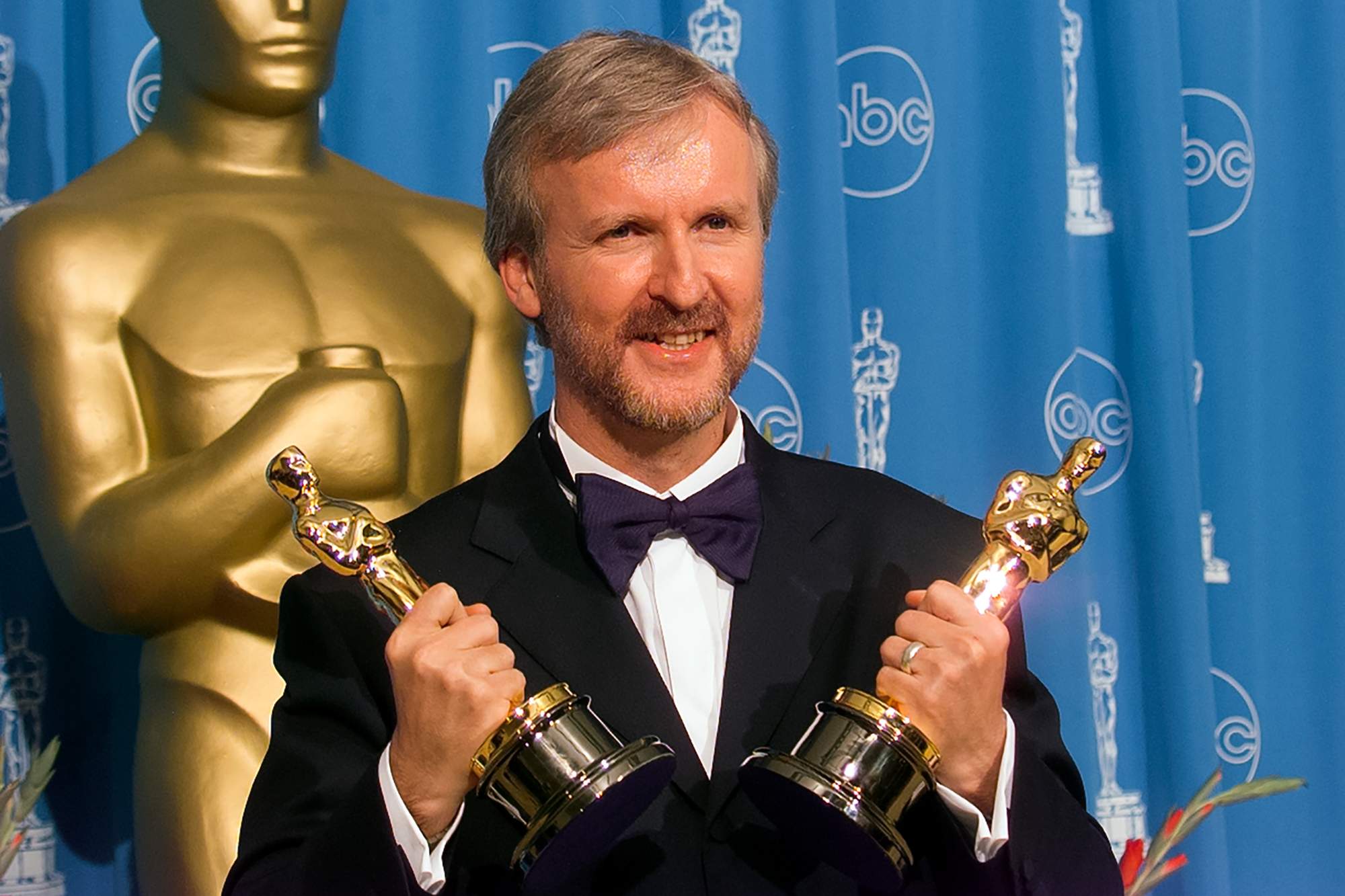 James Cameron, holding two Oscars, which he almost used to hit Harvey Weinstein. He's wearing a tux, standing in front of a blue background and a large version of the Oscar statuette.