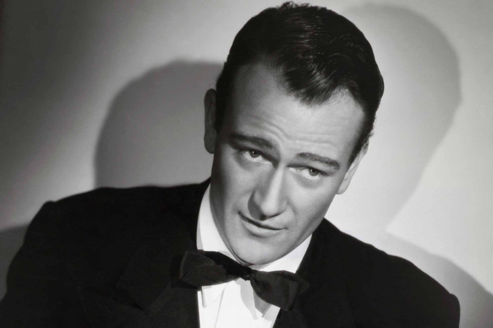 John Wayne, an inspiration to many actors, in a black-and-white portrait photo in a tuxedo.