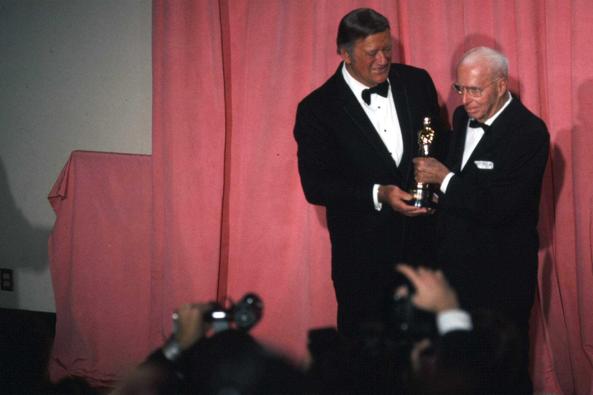 John Wayne and Howard Hawks holding an Oscar together while wearing tuxes
