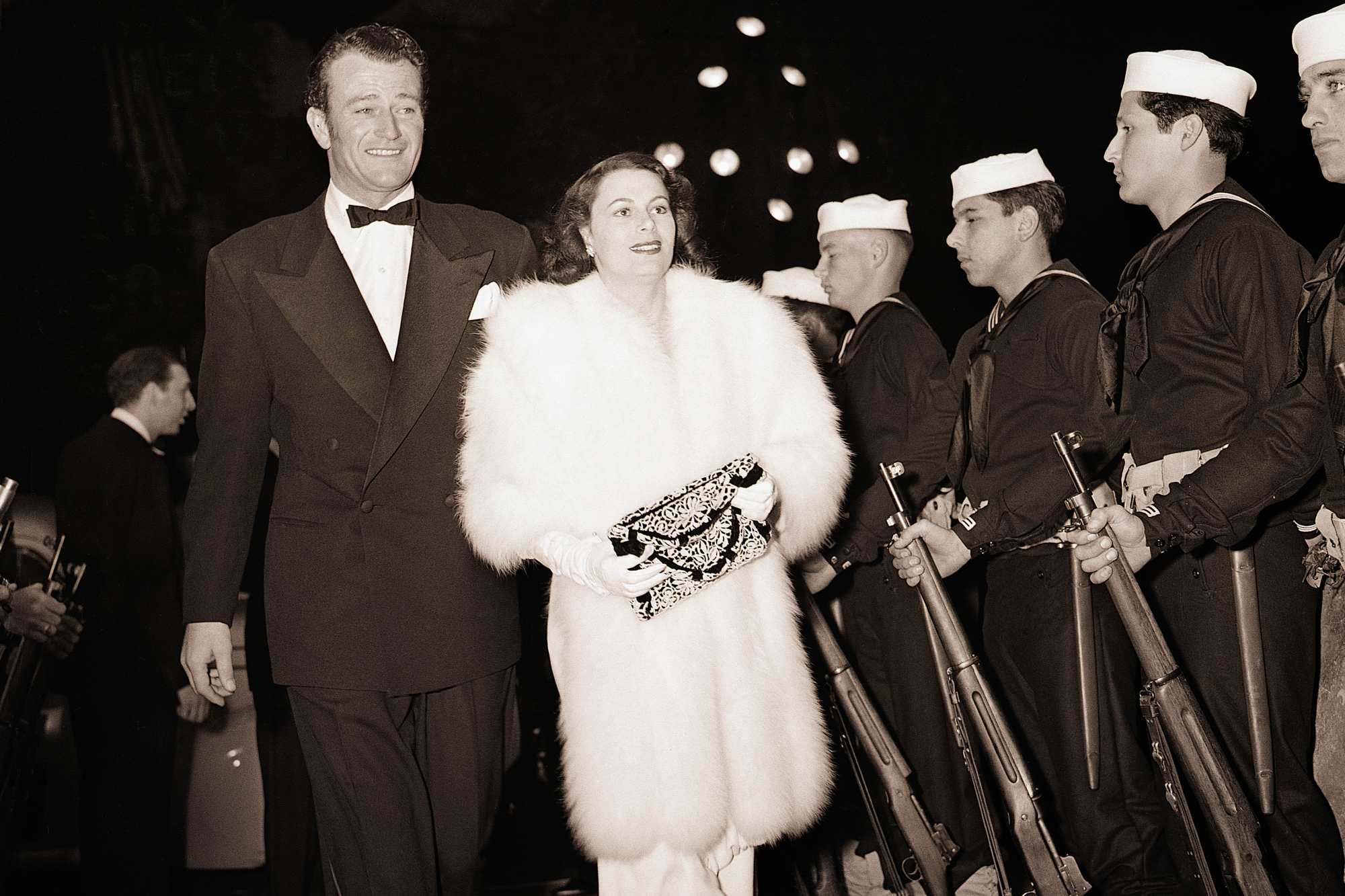 John Wayne and his first wife, Josephine Saenz dressed in formal clothes, walking past a line of men in uniforms.