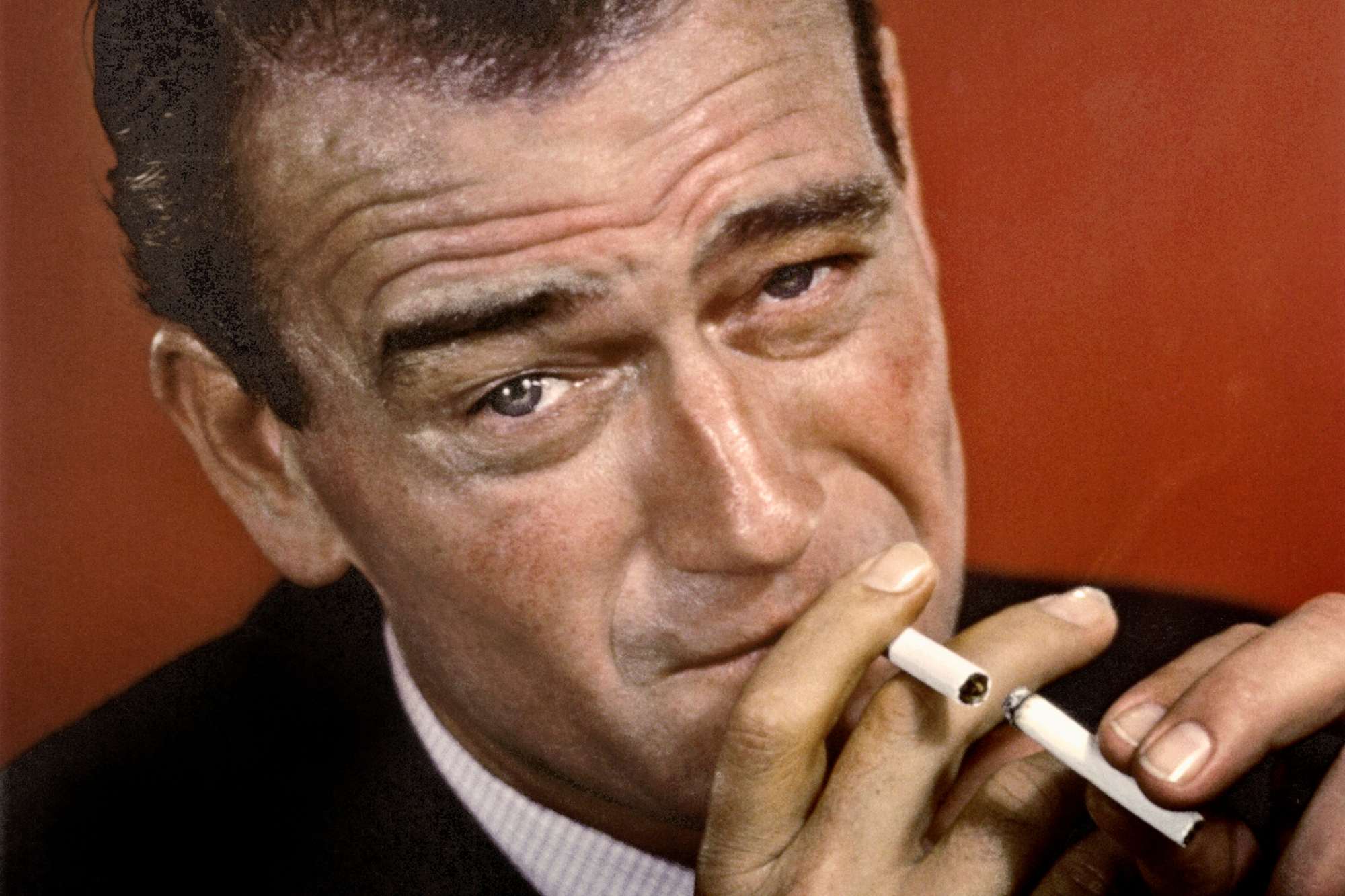 John Wayne image of him lighting his cigarette with another one, looking at the camera in a portrait.