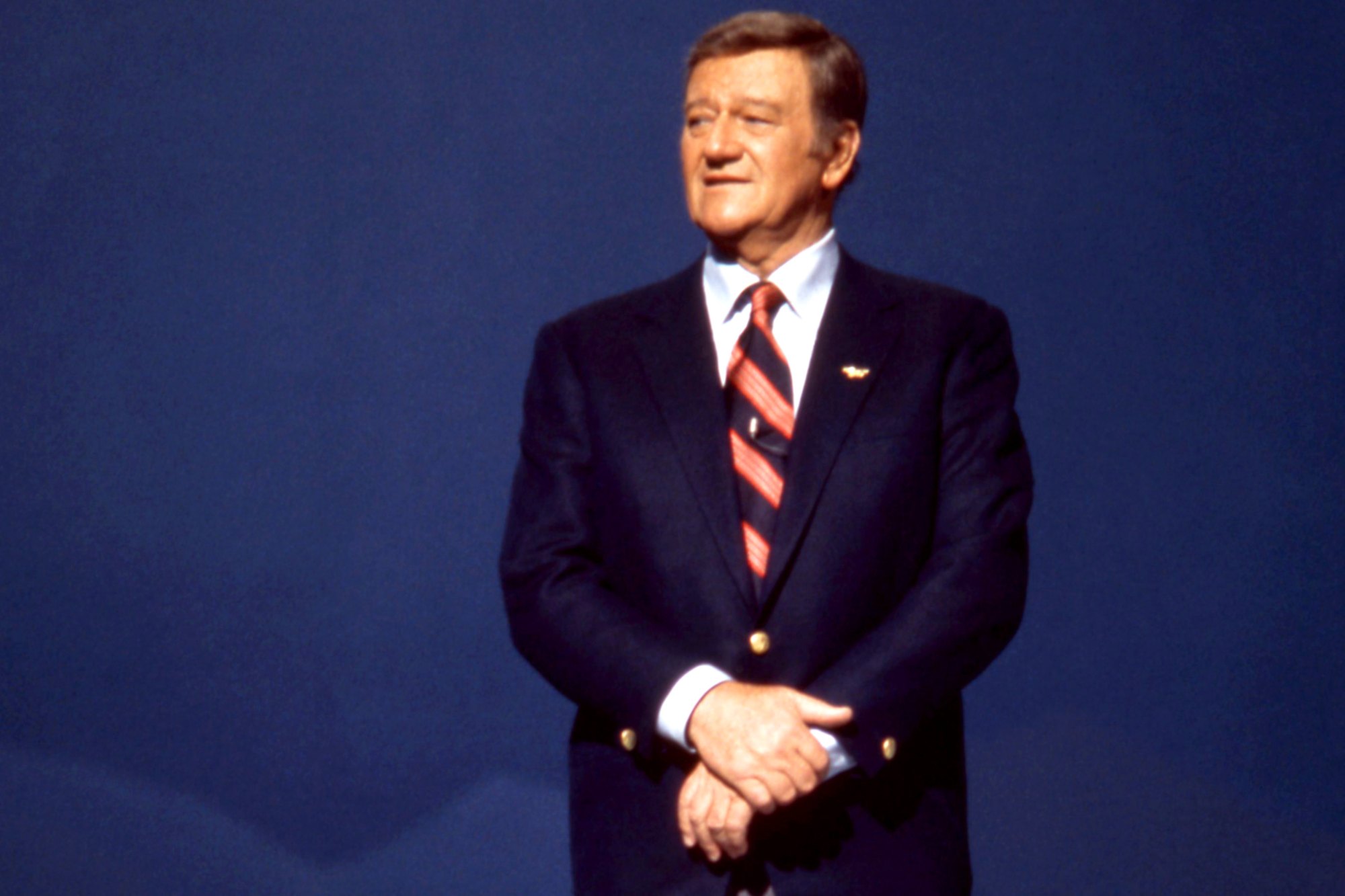 John Wayne, who was a fan of Winston Churchill. He's standing in a suit with his hands crossed in front of a blue background.