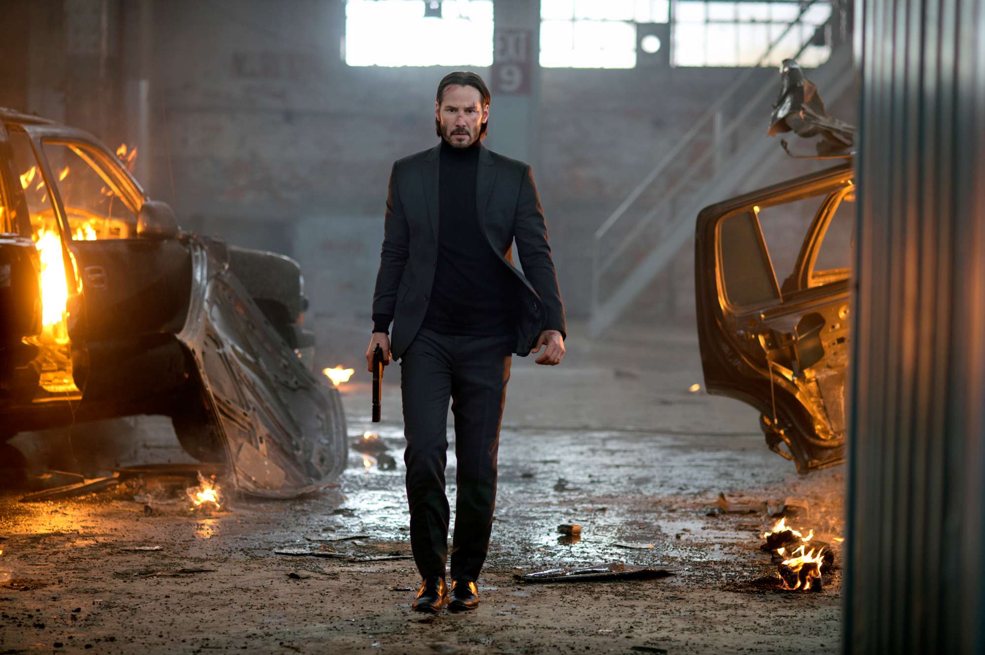 'John Wick' Keanu Reeves as John Wick walking down a path of destruction with fire around him, while he's wearing a suit and holding a gun.