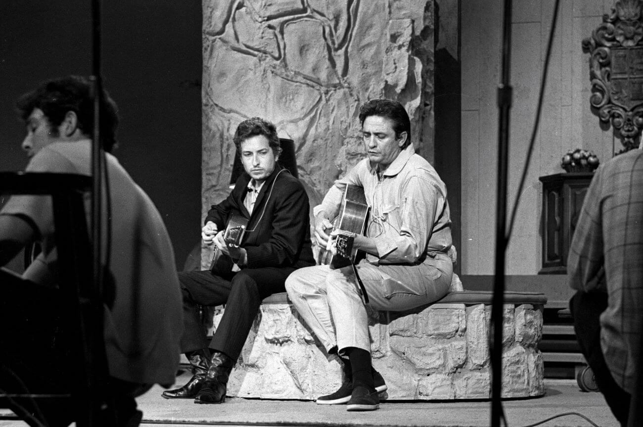 A black and white picture of Bob Dylan and Johnny Cash sitting on a stone bench playing guitars.
