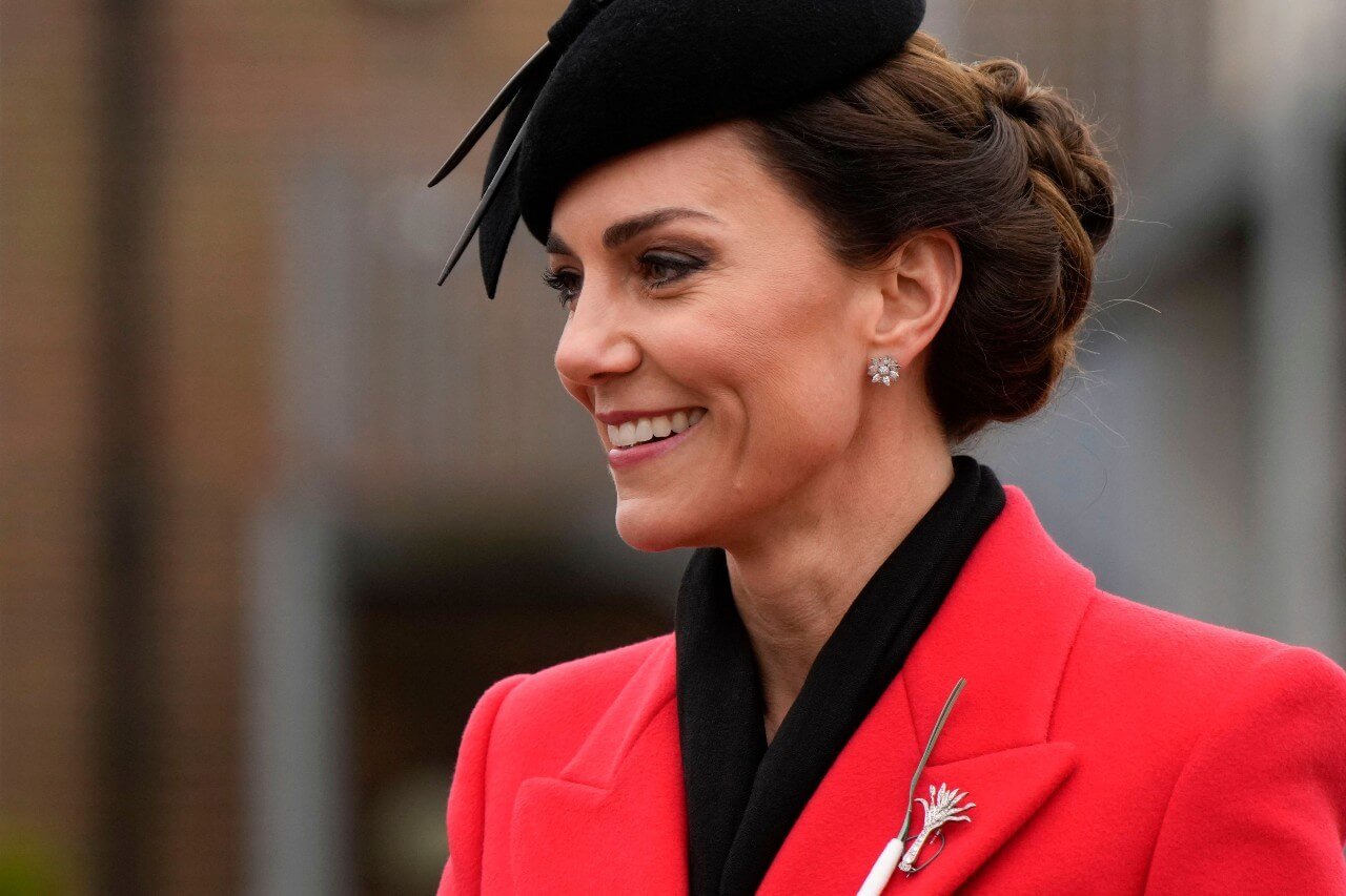 Kate Middleton smiles while wearing a red coat with a diamond brooch and a fascinator.