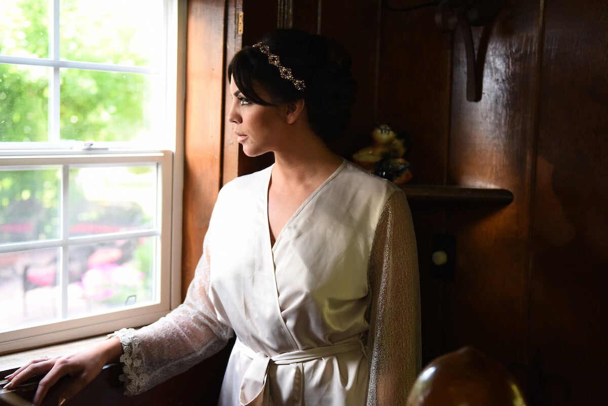 Katie Maloney with makeup and hair done for her wedding in a robe standing next to a window looking out
