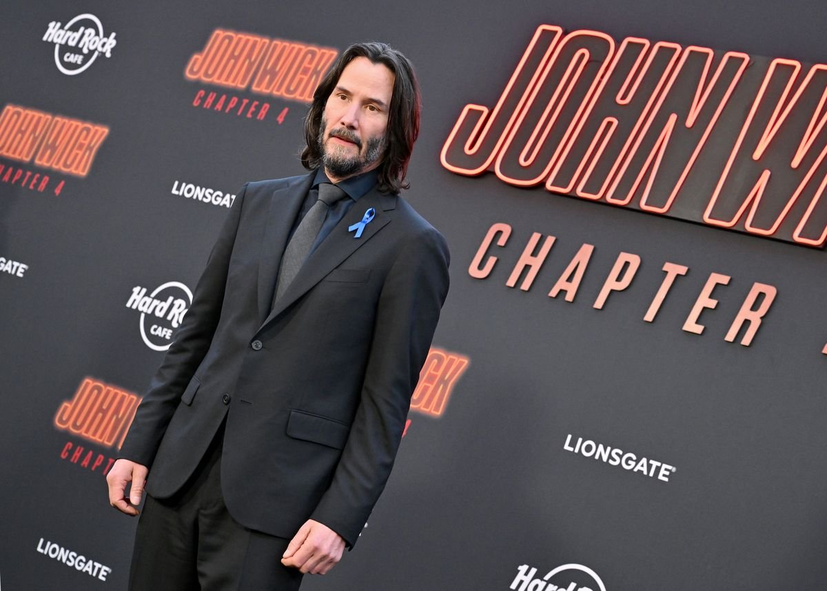 Keanu Reeves poses in front of a black backdrop featuring the "John Wick: Chapter 4" logo.