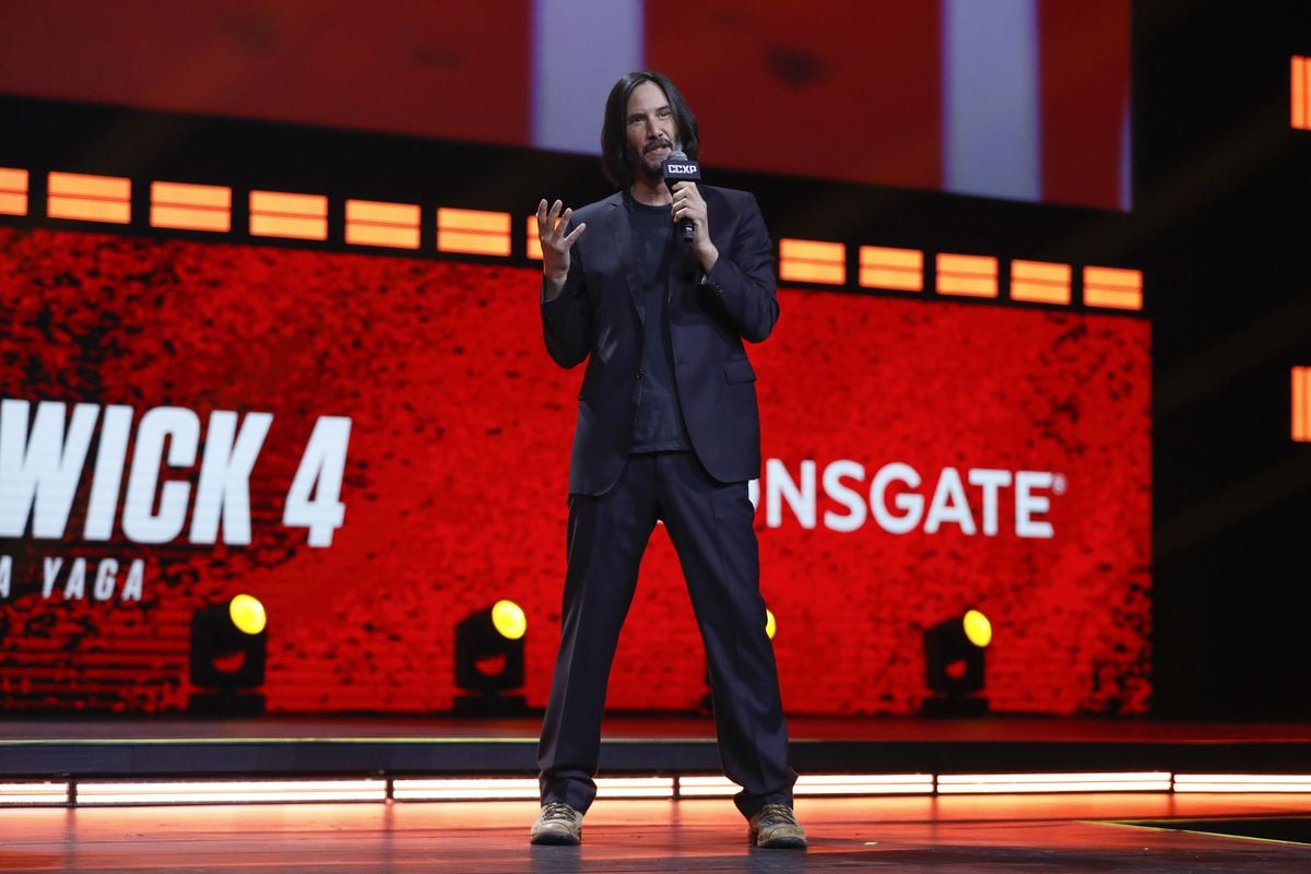 Keanu Reeves stands on stage with a microphone with the "John Wick" logo in the background.
