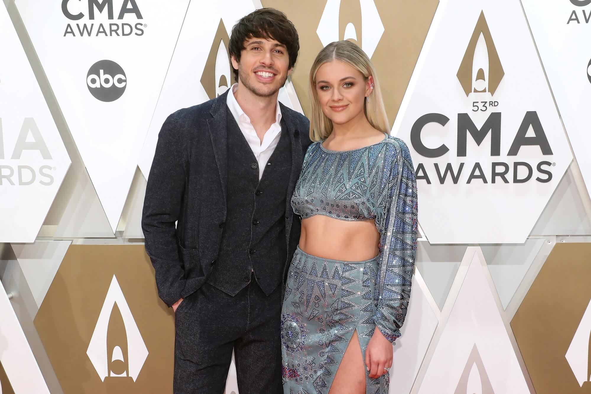 Morgan Evans and Kelsea Ballerini stand together in front of a white and gold CMA Awards backdrop