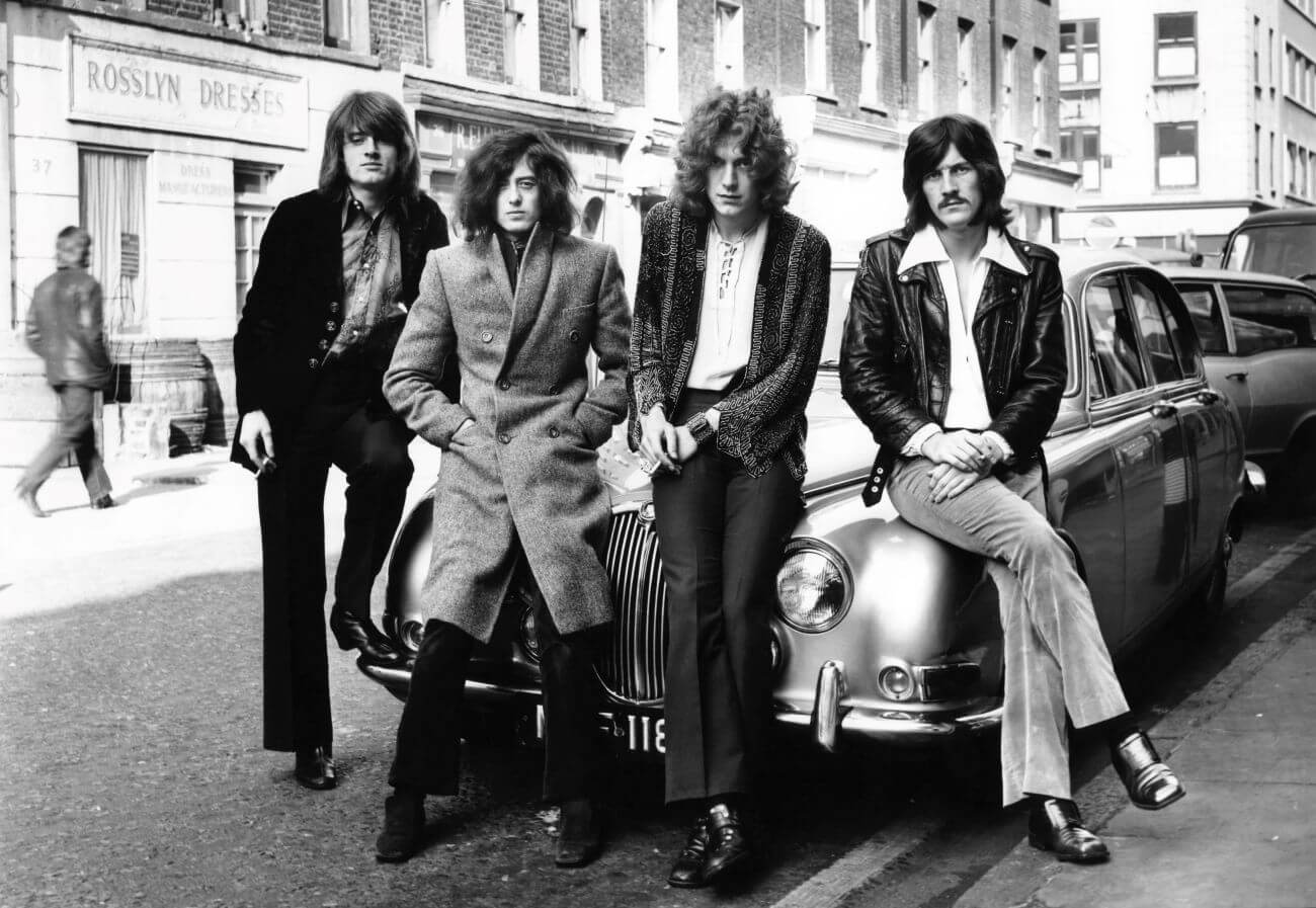 A black and white picture of John Paul Jones, Jimmy Page, Robert Plant and John Bonham of Led Zeppelin sitting on the hood of a car.