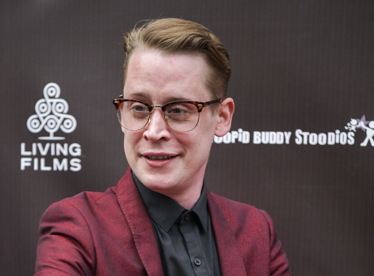 Macaulay Culkin smiles in front of a black background.