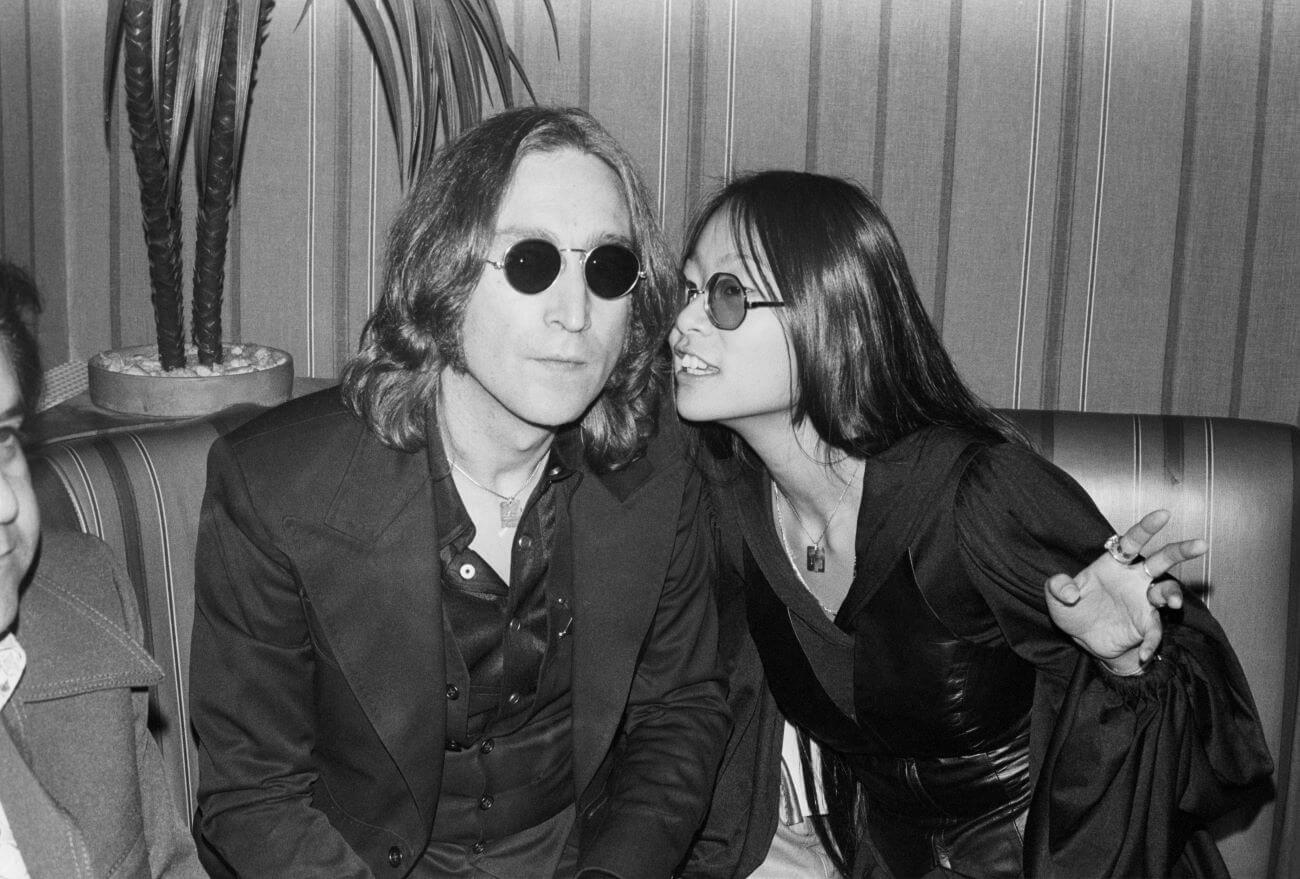 A black and white picture of John Lennon and May Pang wearing sunglasses and sitting on a couch together.