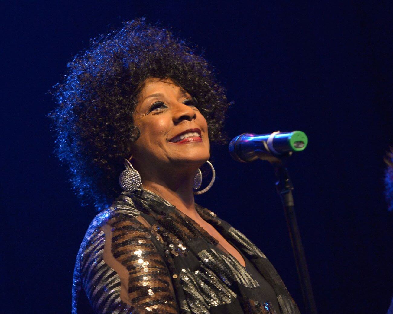 Merry Clayton wears a sequined shirt and stands in front of a microphone.