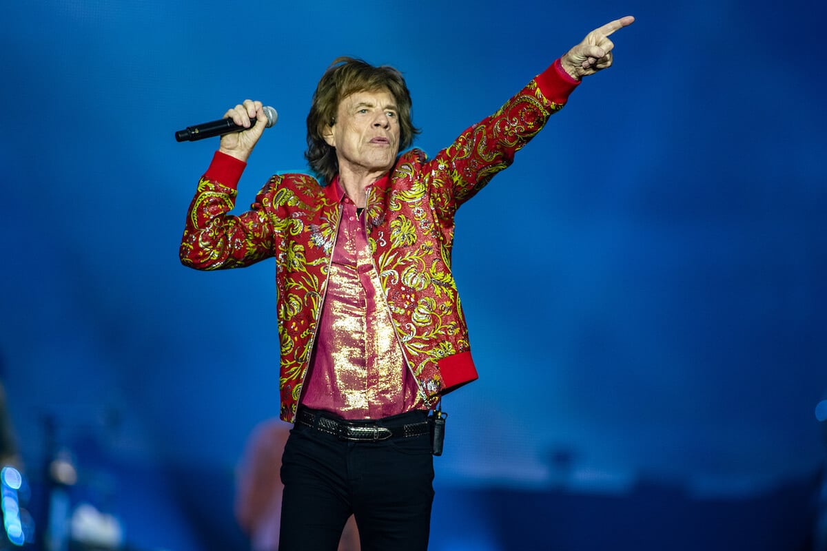 Mick Jagger standing on stage holding a microphone in his right hand and raising is left hand pointing a finger