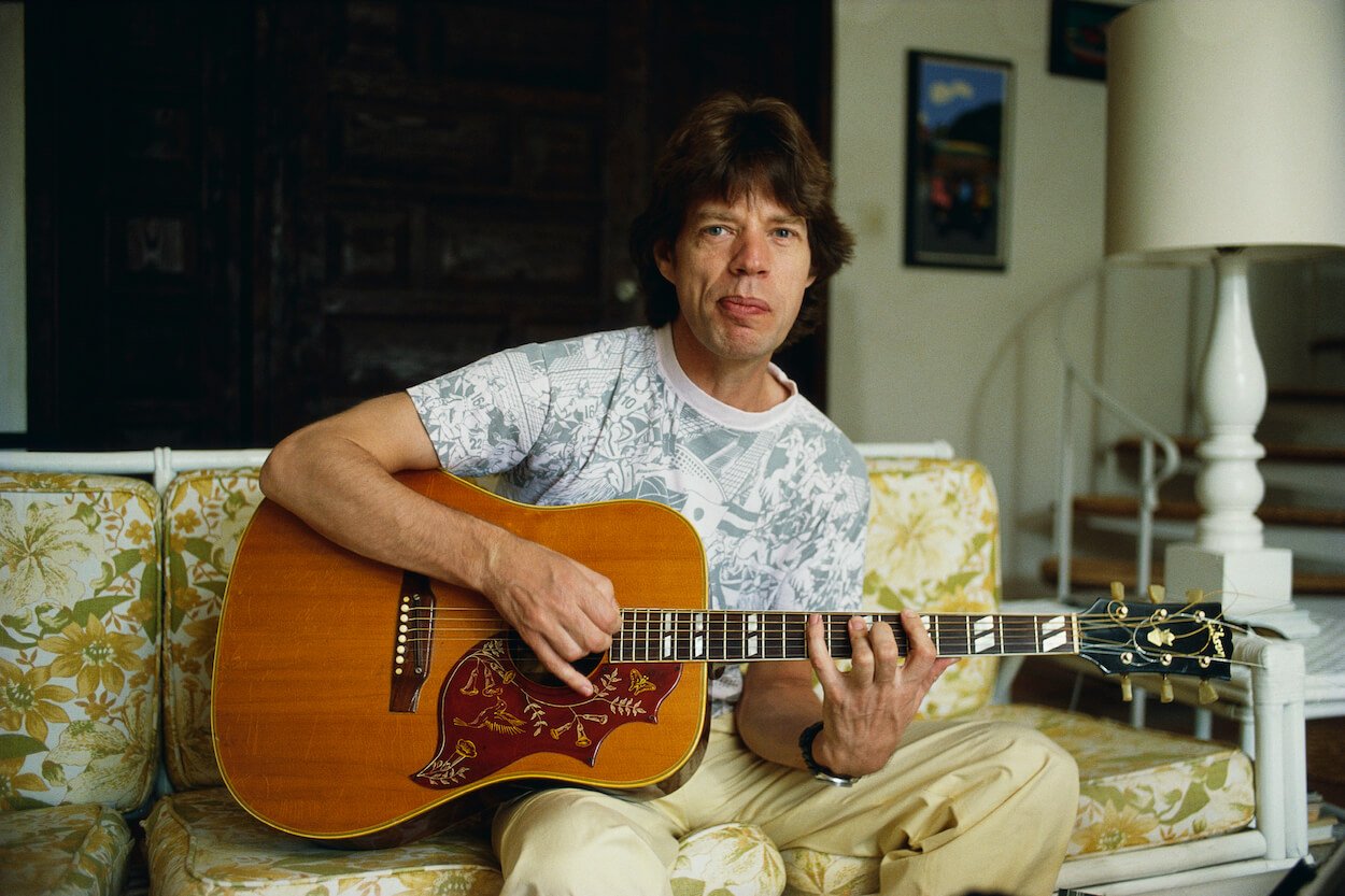 Rolling Stones singer Mick Jagger plays an acoustic guitar while sitting on a couch in 1983.