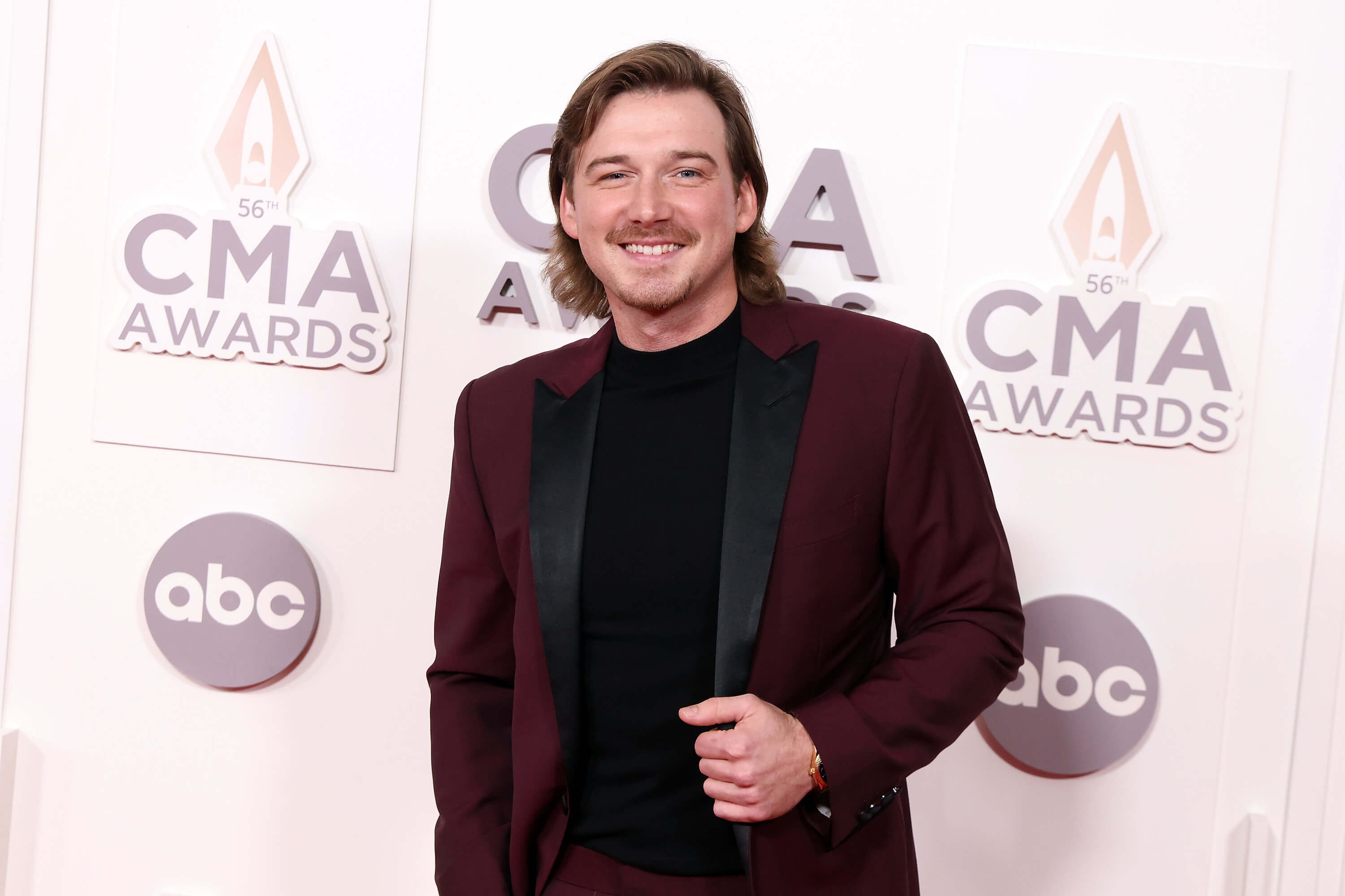 Morgan Wallen stands in front of a backdrop that reads 'CMA Awards' while wearing a burgundy suit