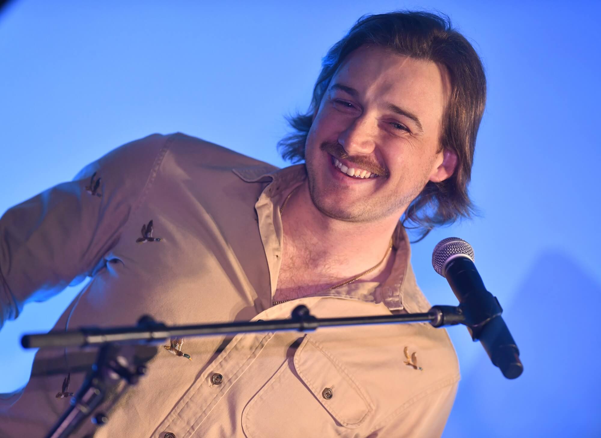 Morgan Wallen smiles in front of a microphone while wearing a brown sihrt