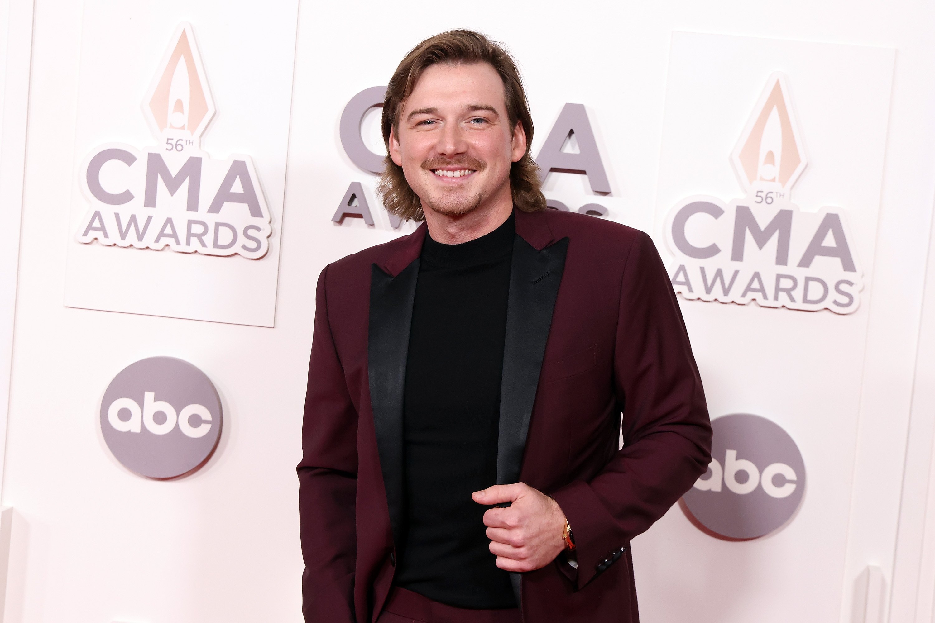 Morgan Wallen stands in front of a white backdrop that reads 'CMA Awards' and 'abc' while wearing a burgandy suit