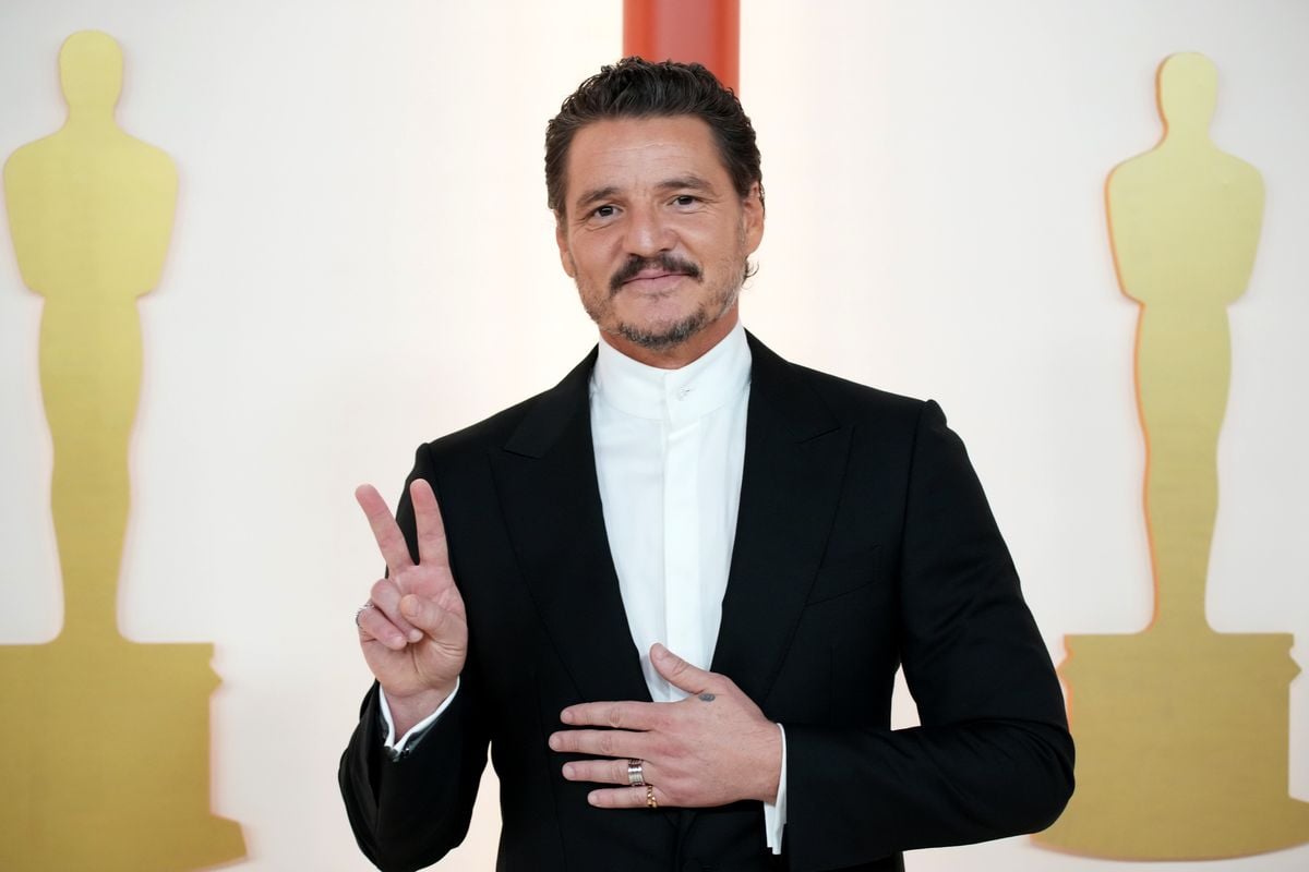 Pedro Pascal makes a peace sign in front of a backdrop featuring the Academy Awards statuette.