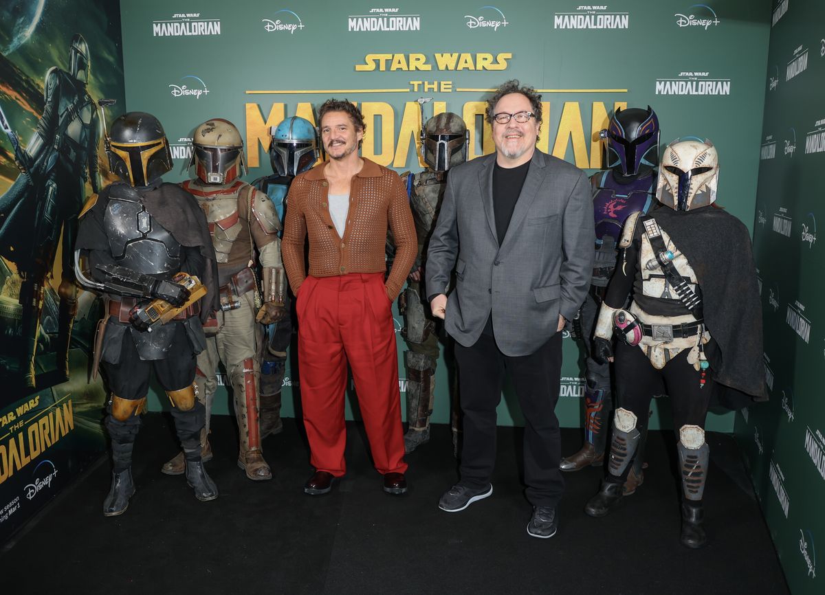 Pedro Pascal and John Favreau pose with several Mandalorians in front of a "The Mandalorian" backdrop.