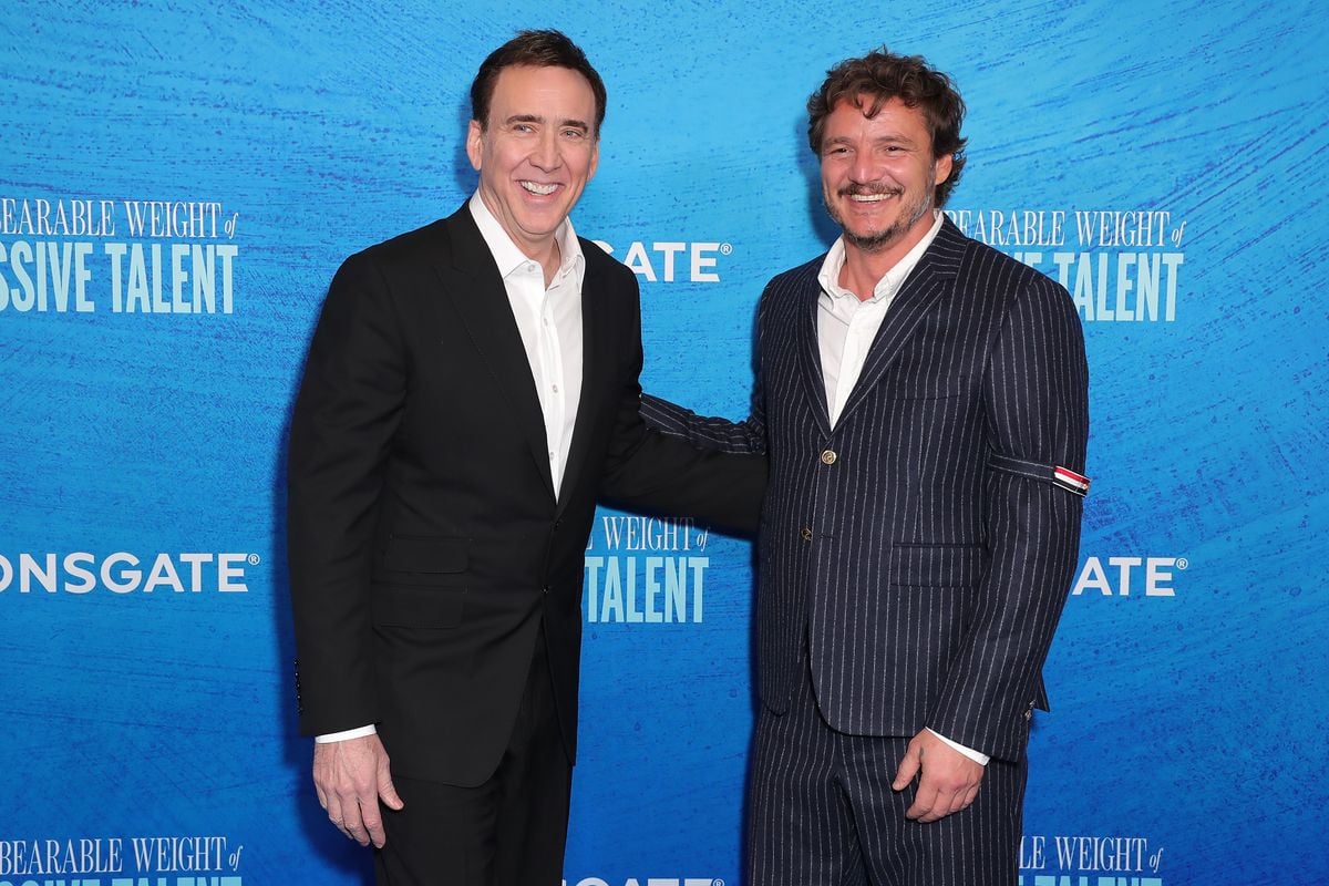 Nicolas Cage and Pedro Pascal pose for photos in front of a blue backdrop featuring the logo for "The Unbearable Weight of Massive Talent"