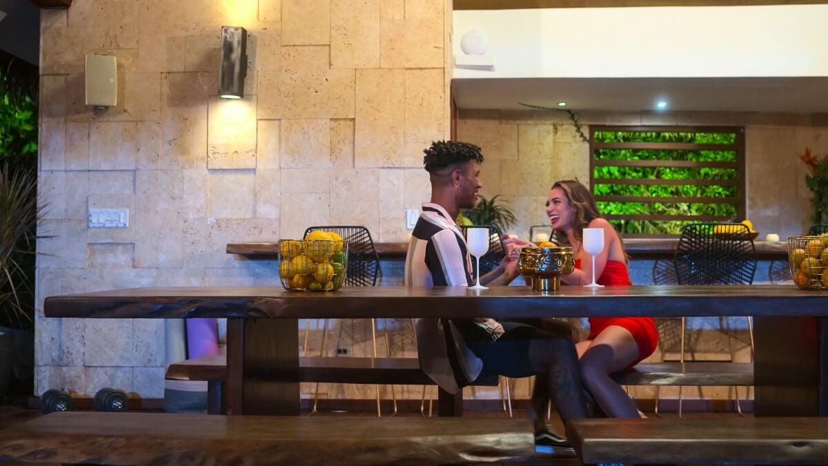 During an episode of Perfect Match, Georgia Hassarati and Dom Gabriel talk while sitting at a table with wine glasses in front of them.