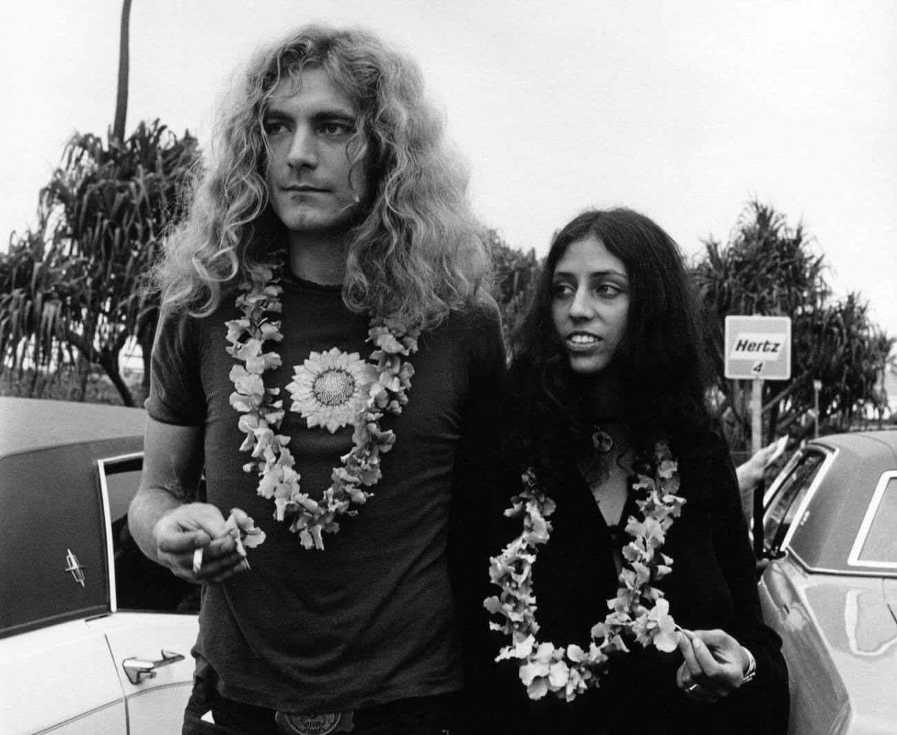 A black and white picture of Robert Plant and Maureen Wilson wearing leis and standing in front of cars.
