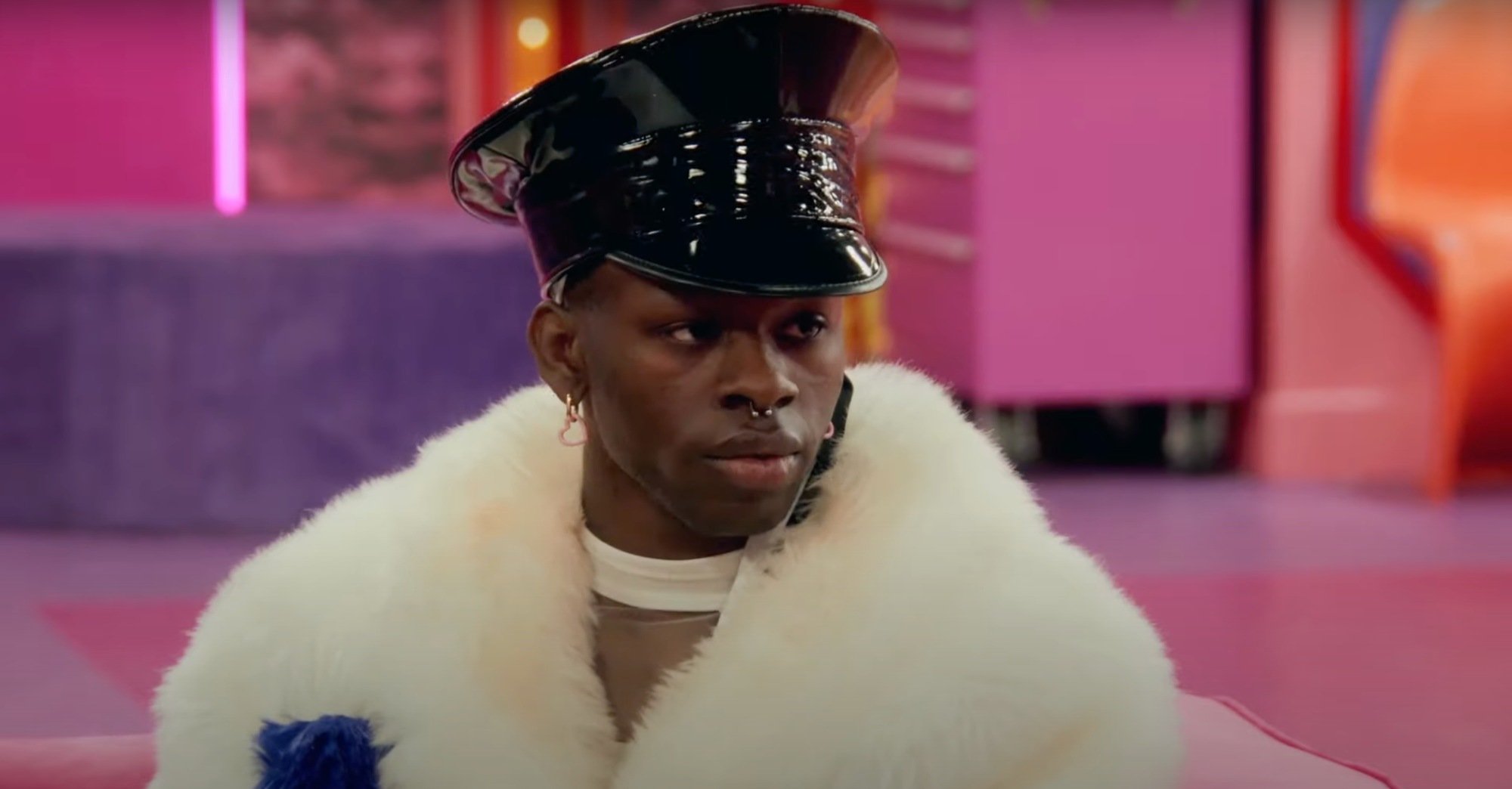 'RuPaul's Drag Race' Season 15 Luxx Noir London in 'Heavengate' looking serious, wearing a black hat and a white fur jacket in the pink Werk room.