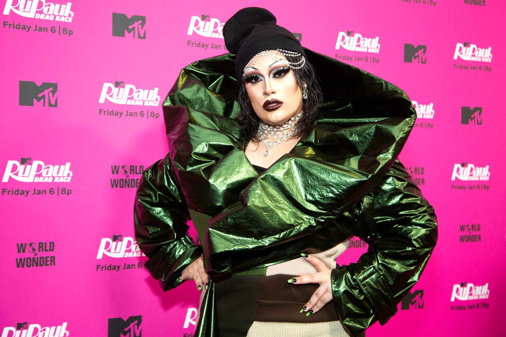 'RuPaul's Drag Race' Season 15 Salina EsTitties wearing a green costume in front of a pink step and repeat. Holding her hands on her hips.