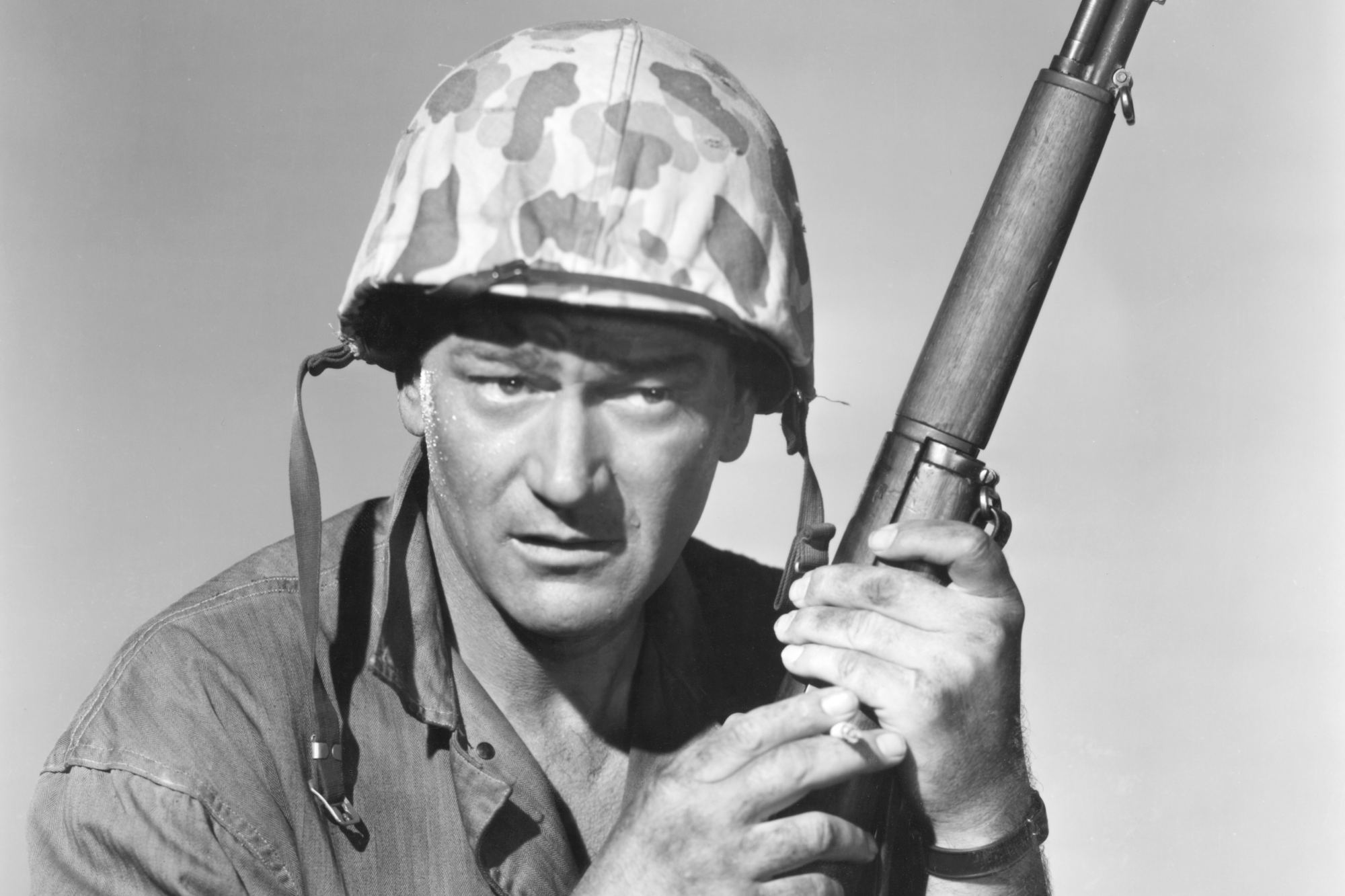 'Sands of Iwo Jima' John Wayne as Sgt. John M. Stryker in war movies. He's holding a gun, wearing his military uniform in a black-and-white picture.