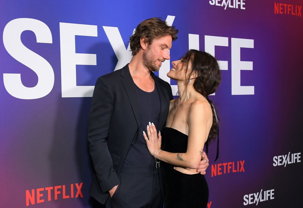 Adam Demos and Sarah Shahi look at each other in front of a "Sex/Life" backdrop.