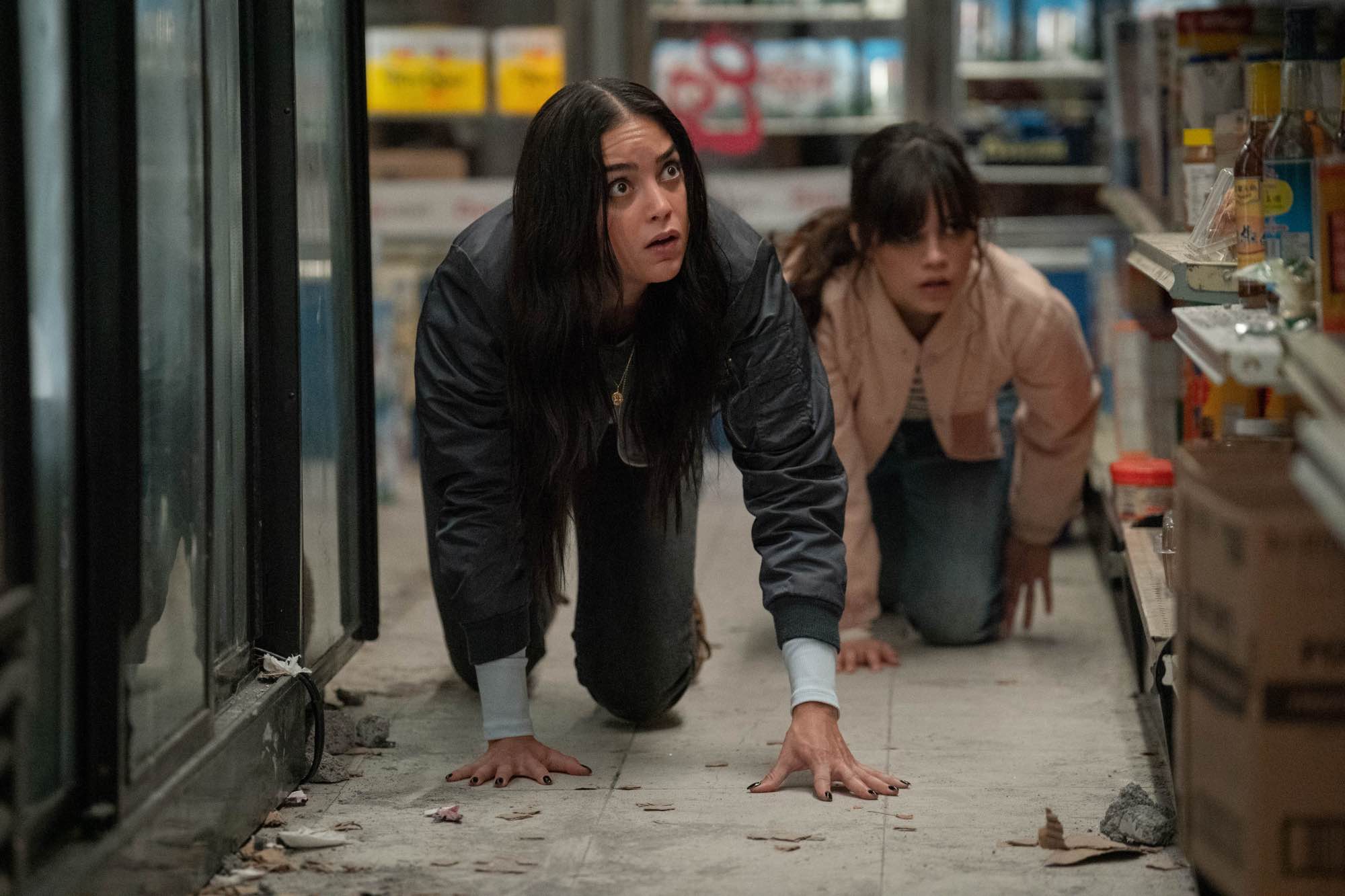 'Scream VI' Melissa Barrera as Sam Carpenter and Jenna Ortega as Tara Carpenter on their hands and knees, crawling on the floor of a bodega looking terrified.