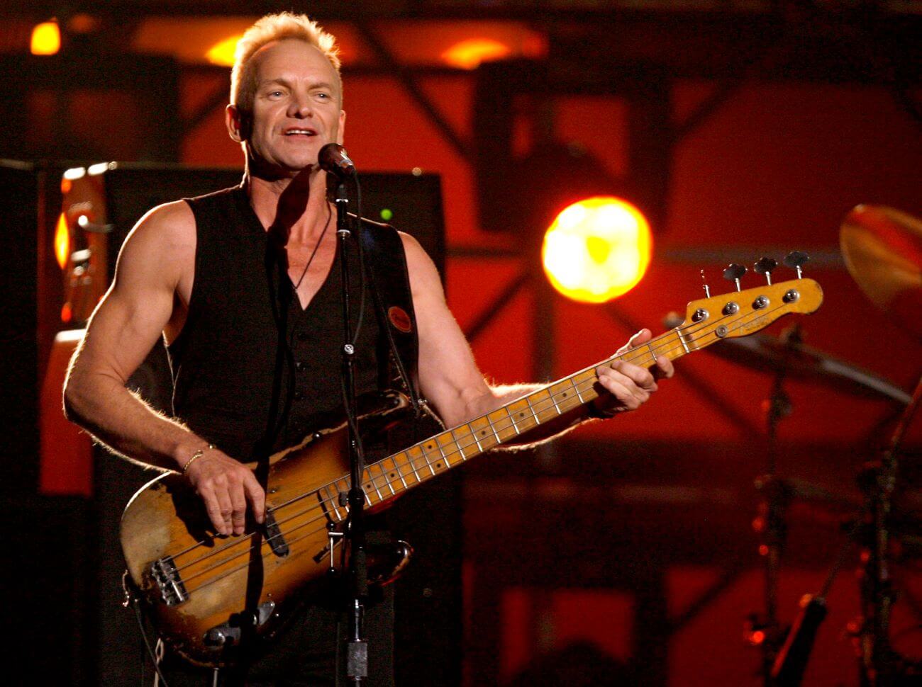 Sting wears a tank top and plays the electric guitar.