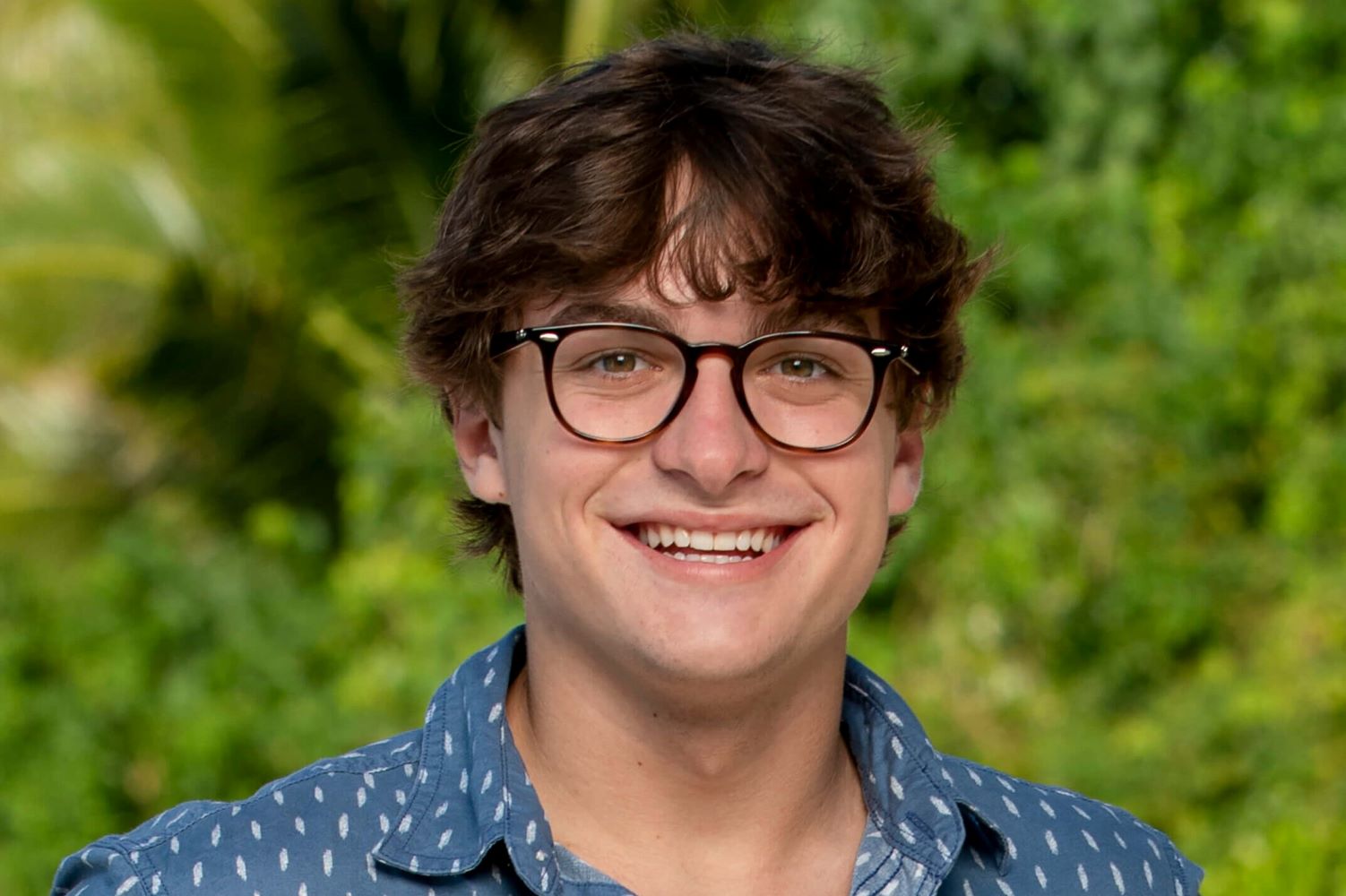 Carson Garrett, who is a part of the 'Survivor 44' cast, wears a blue polo shirt with white dots on it and dark brown-framed glasses.
